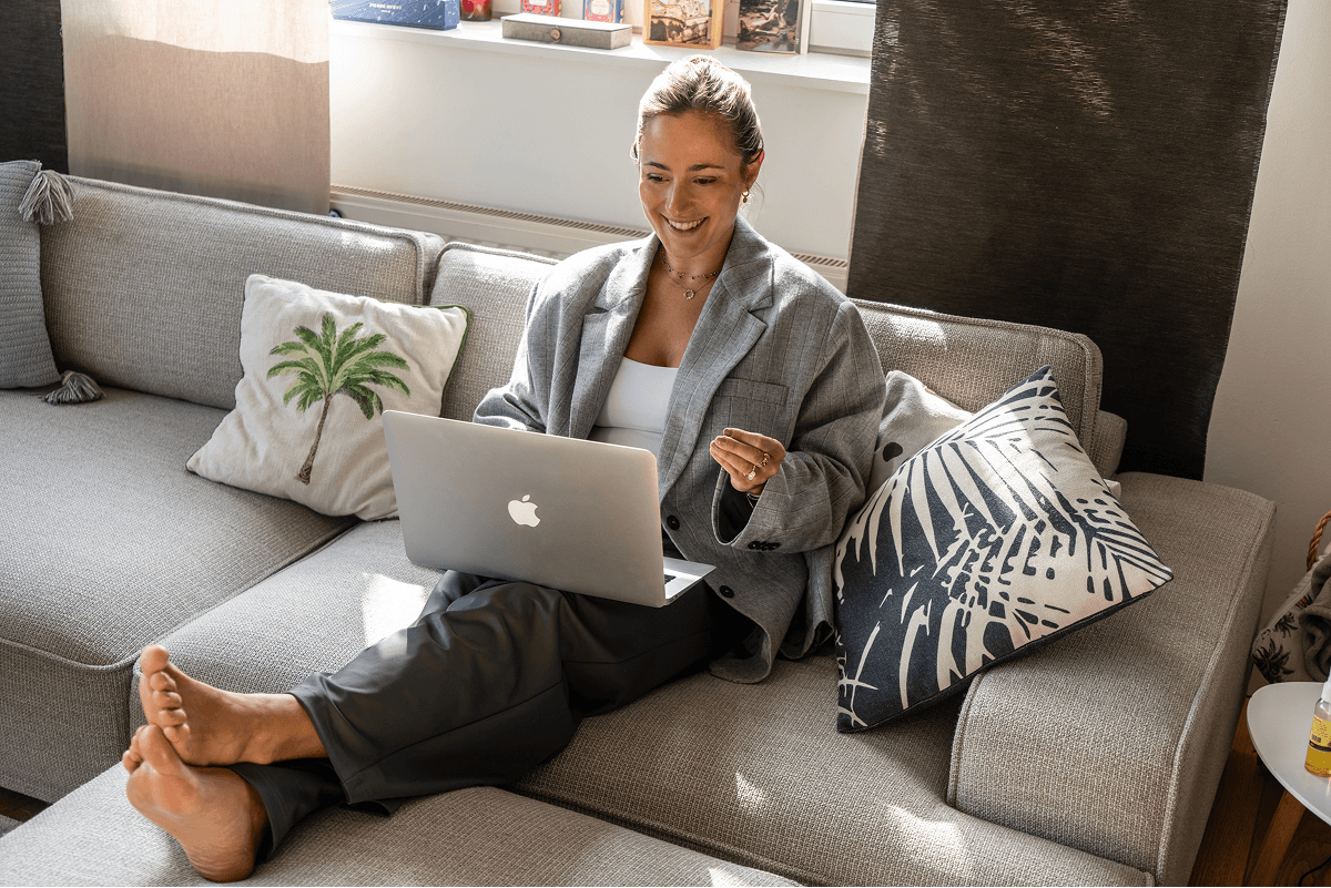 A person is sitting comfortably on a gray sofa, working on a laptop, embodying a relaxed work-life balance in a cozy, sunlit living room with decorative pillows.