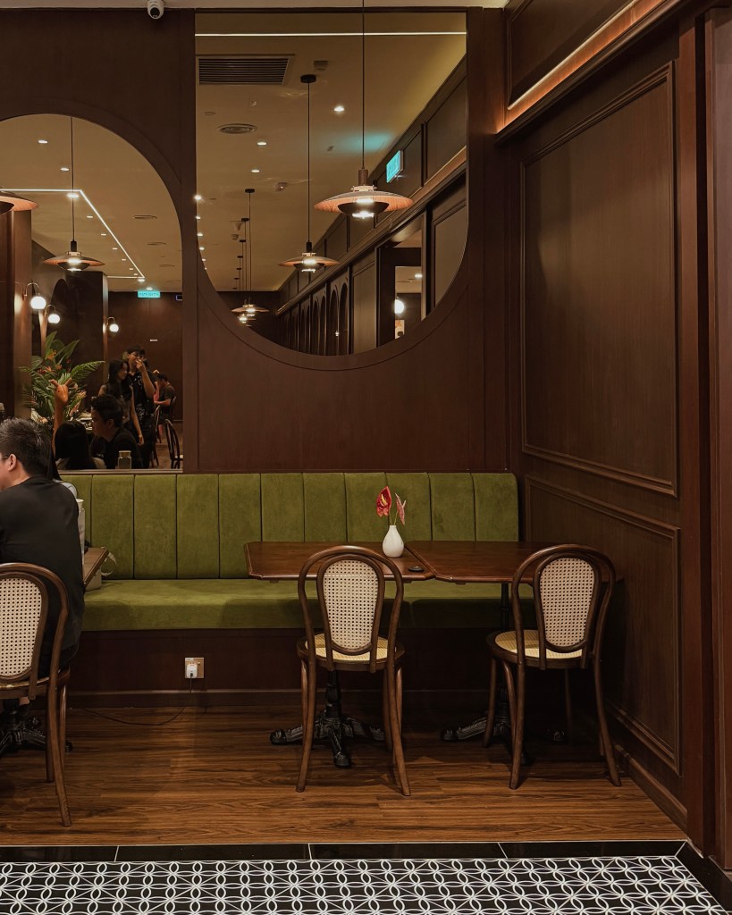 Interior of a restaurant featuring a green bench seat, wooden tables, and stylish chairs. The wall is dark wood, and there are circular mirrors and hanging lights above the tables.