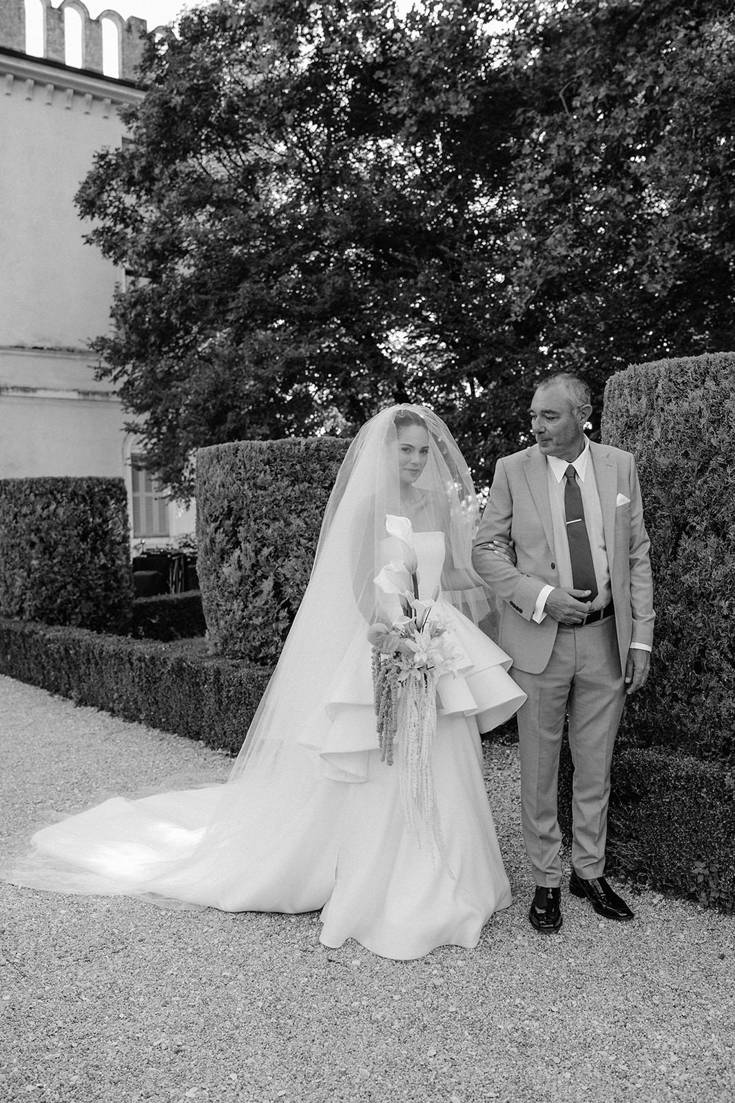 A groom in a tuxedo smiles at a bride in a veil as they exchange vows with an officiant in a black suit.