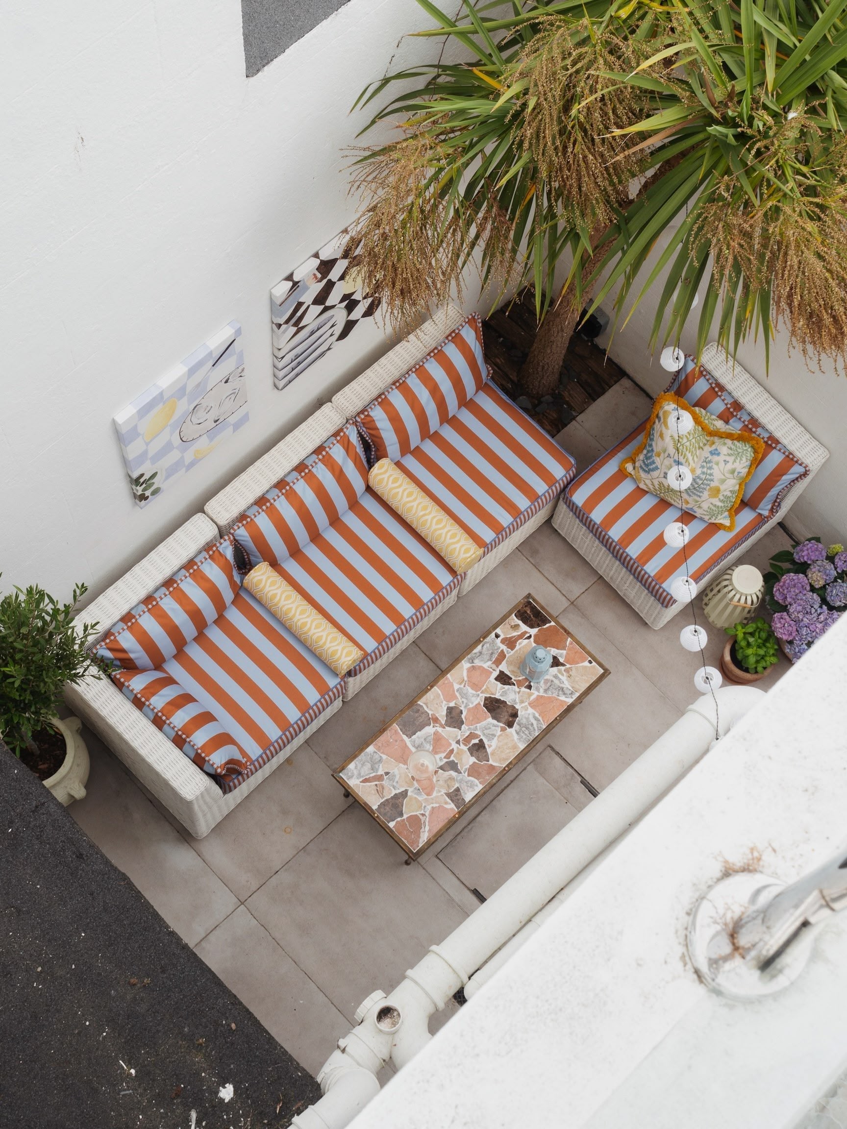 View of a small outdoor balcony with a striped orange and white sofa, a matching armchair, a mosaic coffee table, hanging string lights, a large potted palm, purple hydrangea flowers, and wall art, from a top-down perspective.