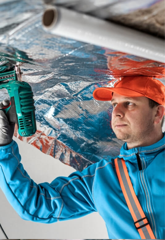 A person in a blue jacket and orange hard hat uses a power tool on a ceiling, focused on their task.