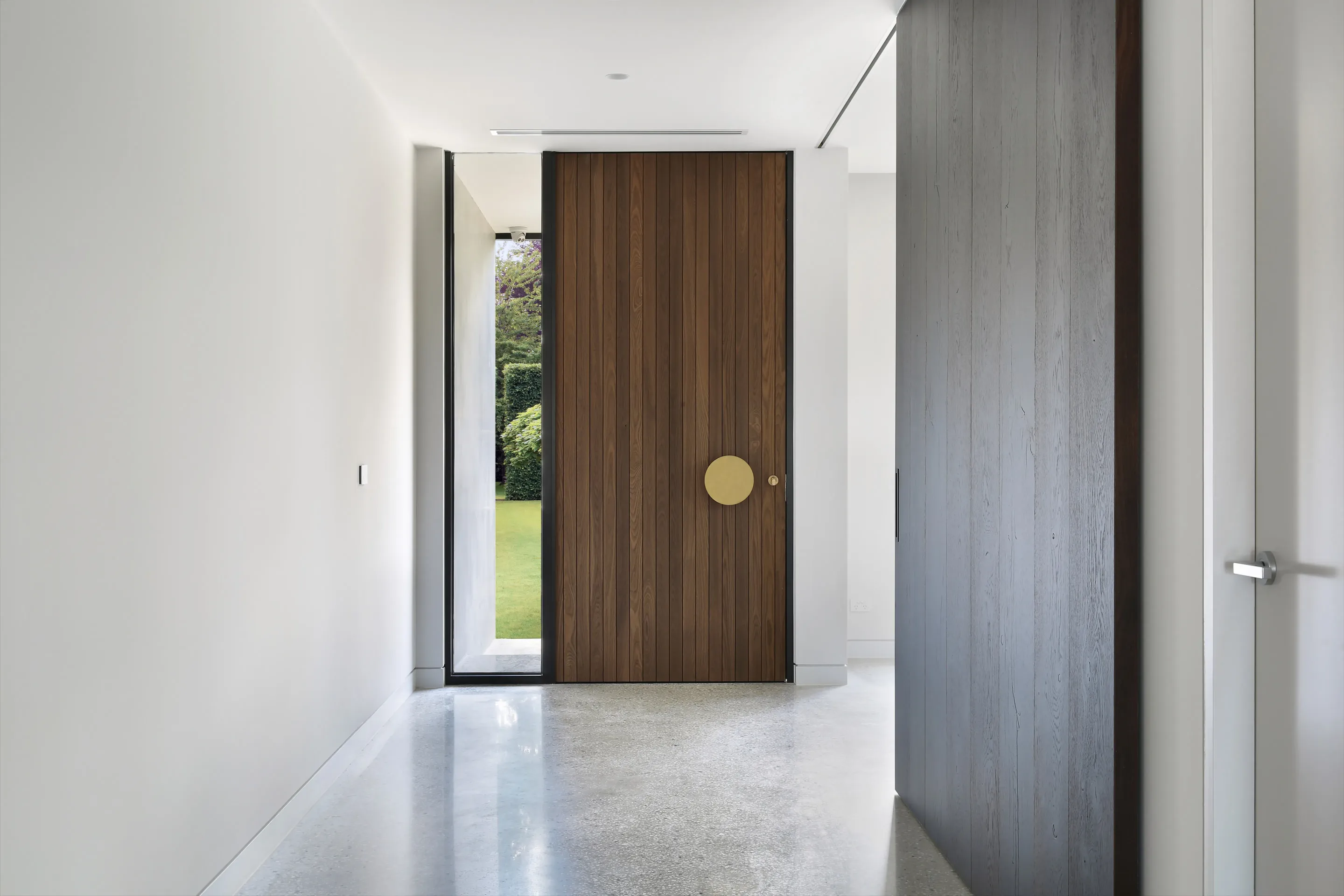 Entry hallway with white walls, polished-concrete floor, a timber-batten pivot door with brass handle and sidelight, and a dark timber-clad feature wall to the right