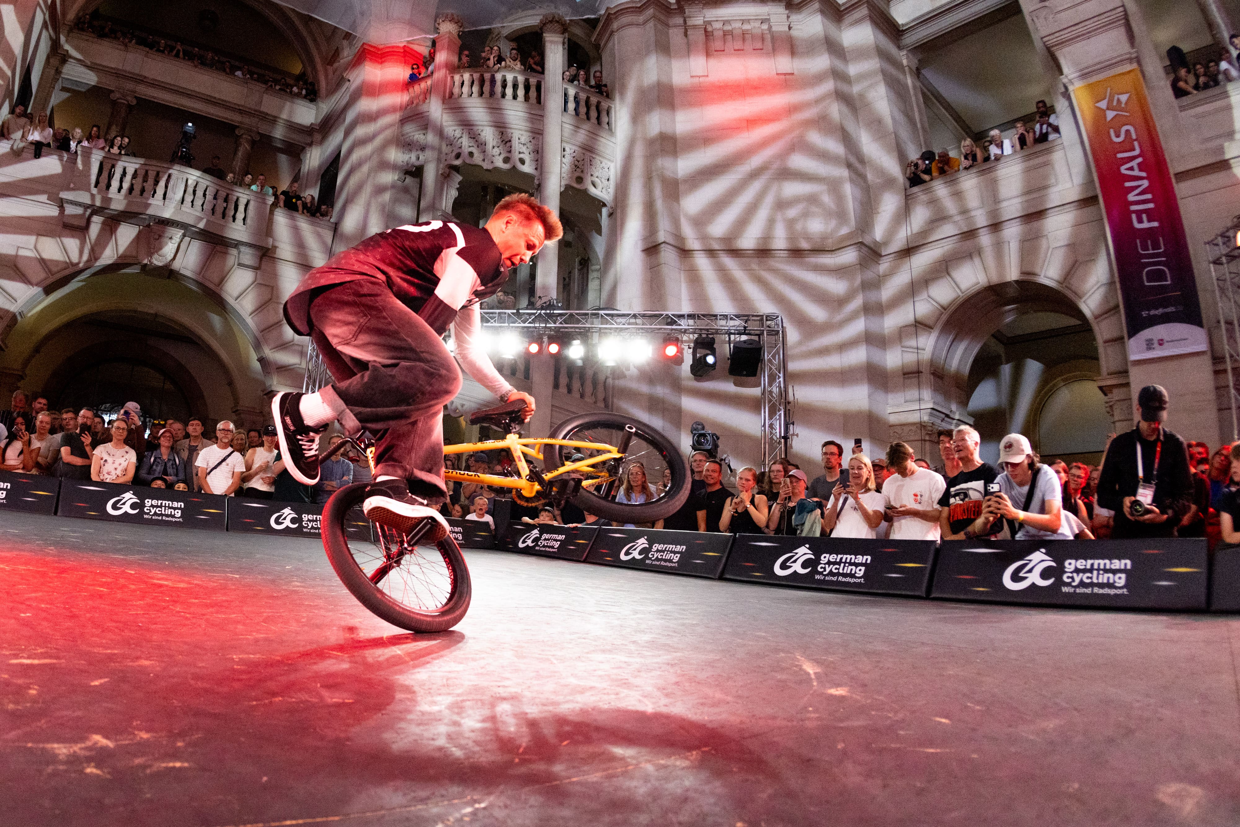 Chris Böhm macht einen BMX-Flatland-Trick auf dem Hinterrad bei den BMX Finals in einer prächtigen Halle, gelbes Bike, rotes Licht und klatschendes Publikum im Hintergrund