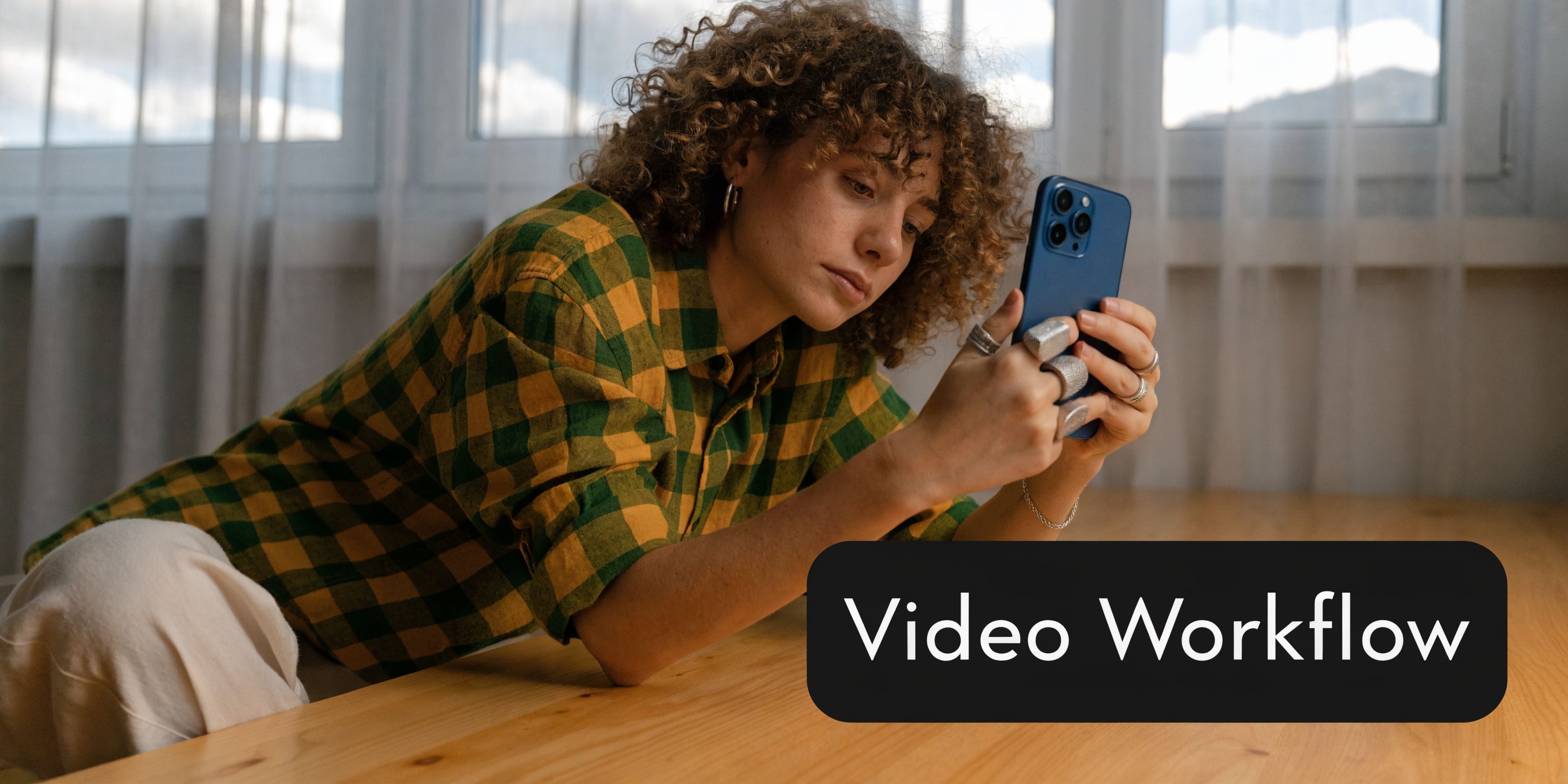 A young woman with curly hair sits at a wooden table while looking at her smartphone.