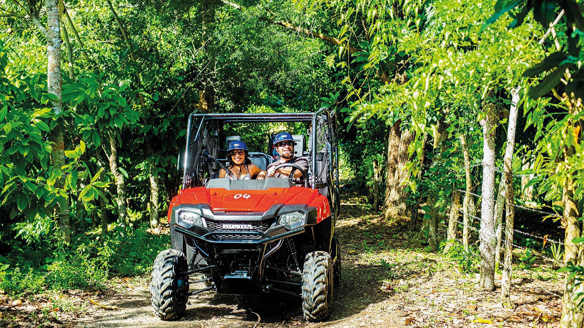 Two tourists driving a red off-road buggy on a dirt trail through a lush green jungle.