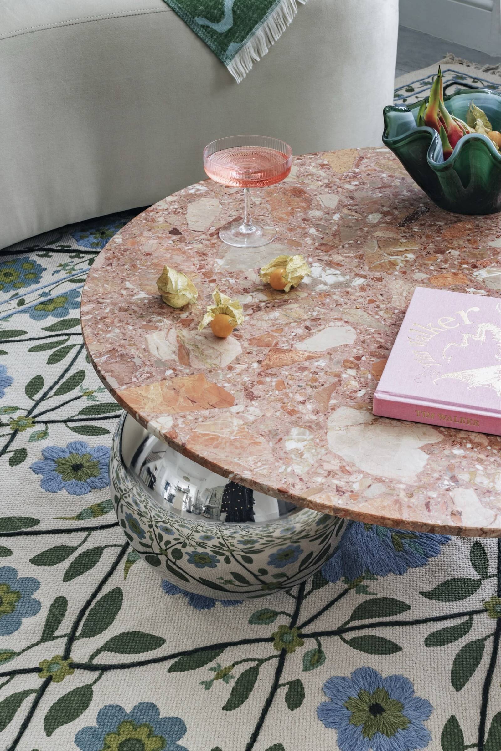 Round pink marble table with a pink cocktail, dried physalis fruit, a pink book, and a green vase with colorful flowers, on a floral rug in a living room.