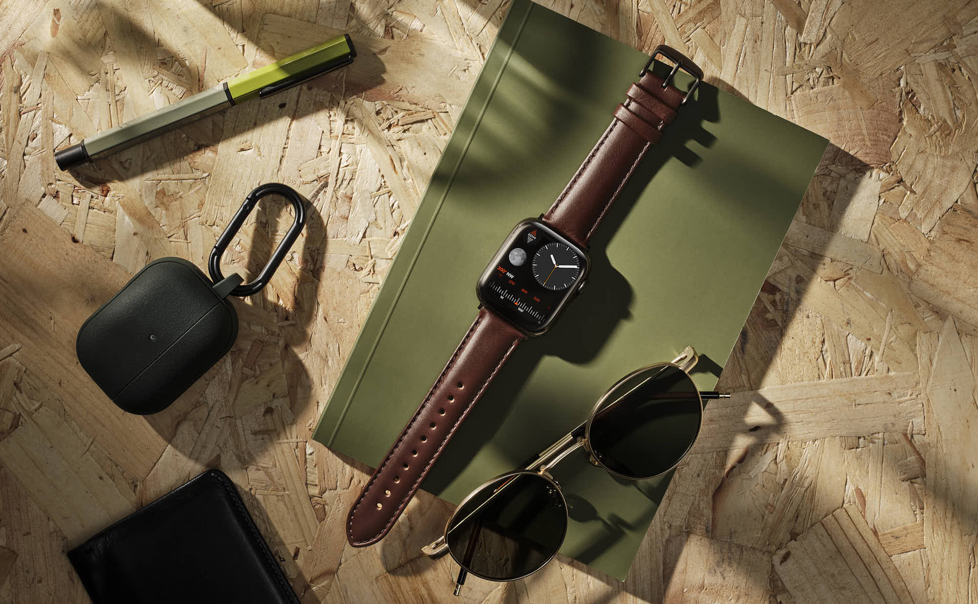 Smartwatch with leather strap, stylish sunglasses, pen, and wireless earbud case on cork surface.