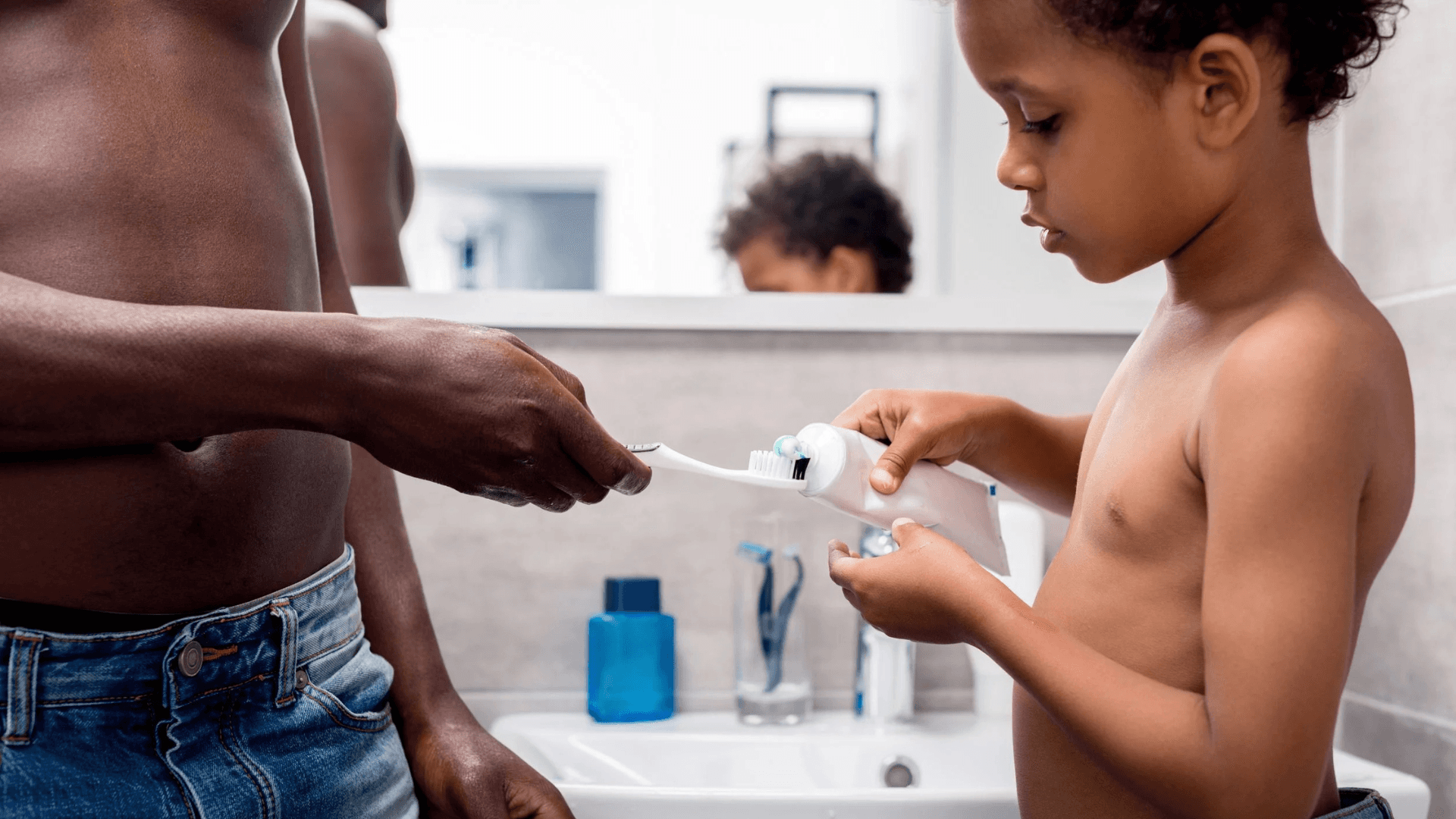 Adult helping a young child put toothpaste on a toothbrush at a bathroom sink