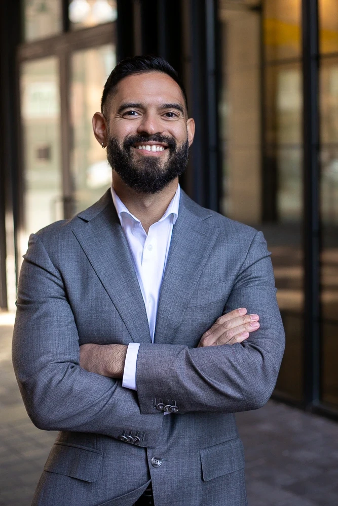 Um indivíduo confiante com barba e braços cruzados, vestindo um terno cinza e camisa branca, está em um ambiente urbano com janelas de vidro ao fundo.