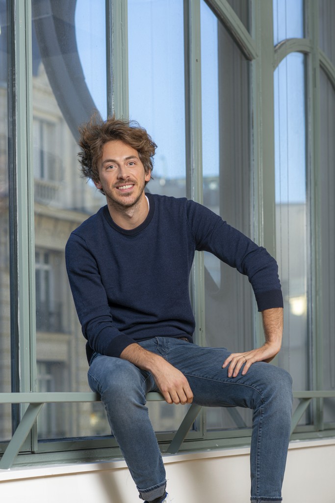 Young person with wavy, shoulder-length red hair, wearing a rust-colored vest over a beige shirt. A brown scarf is tied neatly at the neck. Neutral backdrop.