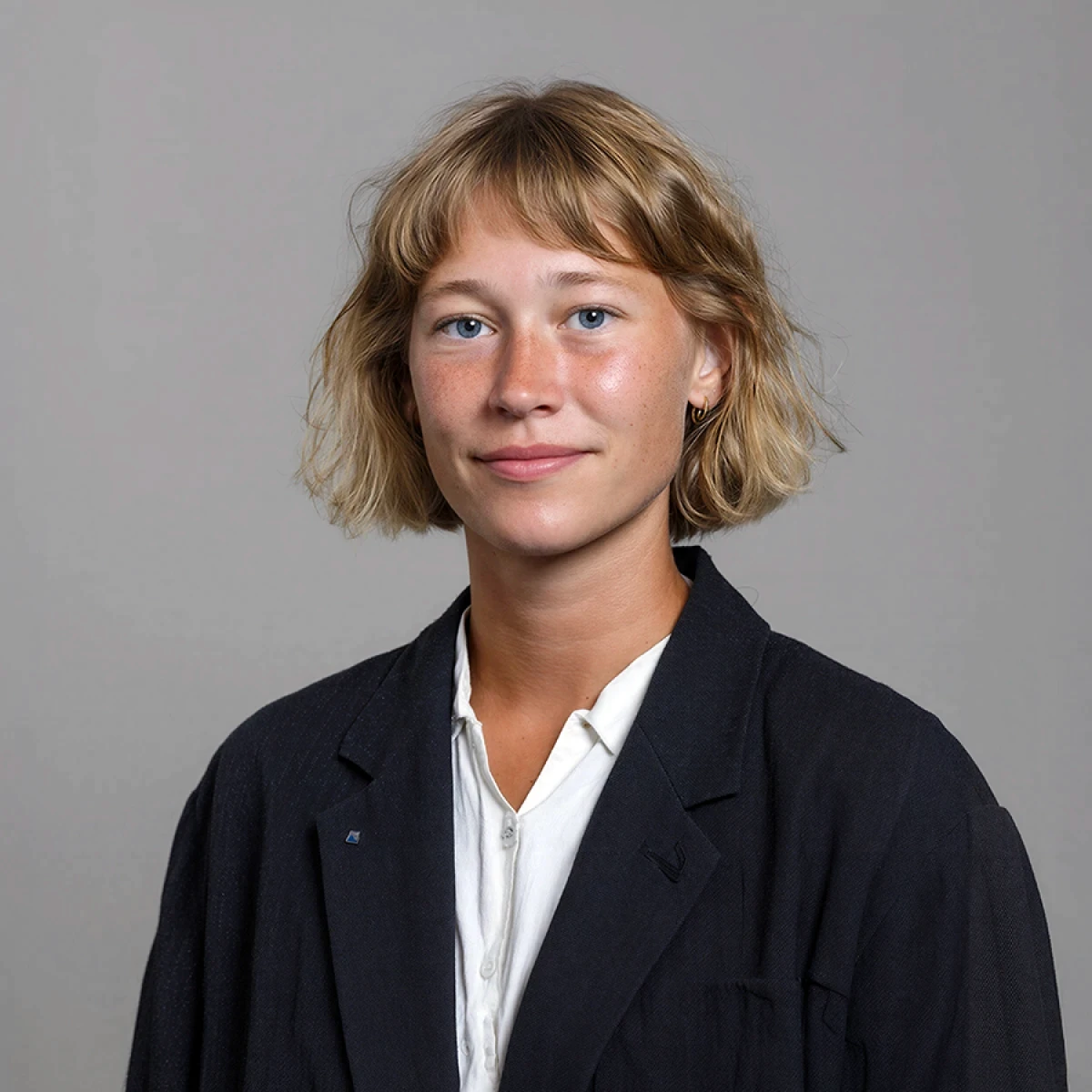 A person with short, wavy hair and a black jacket smiles against a neutral gray background.