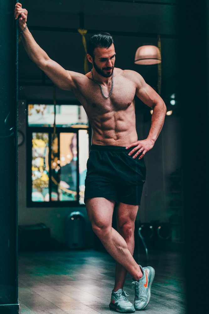 A man with defined abs holding onto a vertical pole in a gym, looking to the side. 