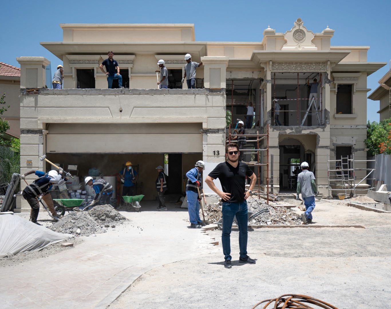 A group of construction workers in Jumeirah Islands, wearing helmets and safety gear, are busy renovating the facade and landscaping of a two-story house with ornate architectural details and a large garage.