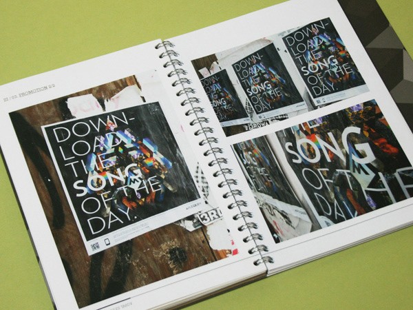 An open spiral-bound book displays pages with text and images. The left page features the text "DOWNLOAD THE SONG OF THE DAY" on a black background with colorful, graffiti-style artwork. The right page shows smaller images of similar artwork and text, including “DOWNLOAD THE SONG OF THE DAY” repeated on multiple pages. The book rests on a green surface.