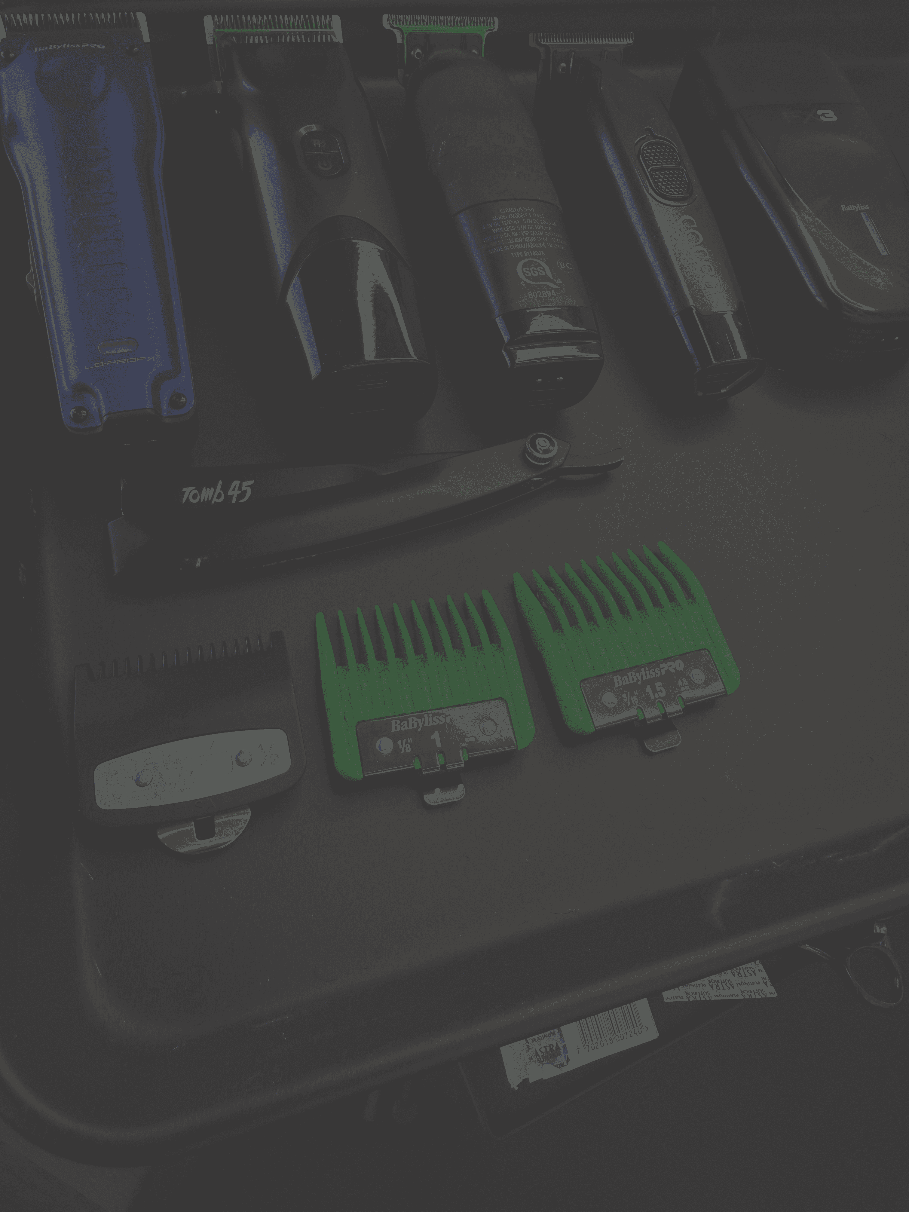 Close-up of barber tools on a workstation, including clippers, trimmers, a straight razor, green clipper guards, and other grooming accessories.