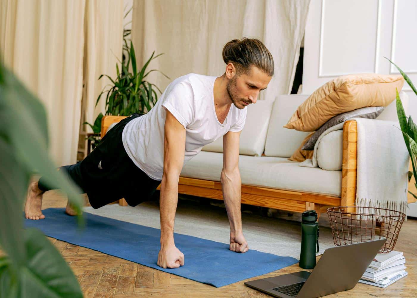 Man on a blue yoga mat doing a plank on his fists while staring at open laptop in front of him