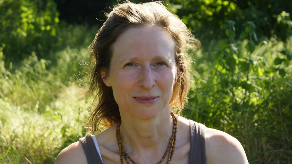 Yogalehrerin (Karina Gaurangi Schmuck) mittleren Alters mit hellbraunen Haaren und einem leichten Lächlen, mit Holzketten und einem grauen Top ist bis zum Schlüsselbein vor einer verschwommenen wilden Wiese zu sehen