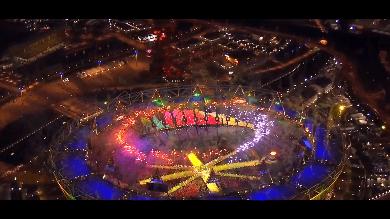 Aerial view of a brightly lit stadium during a night event, showcasing vibrant multicolored light patterns across the field, surrounded by a large cheering crowd and city lights.