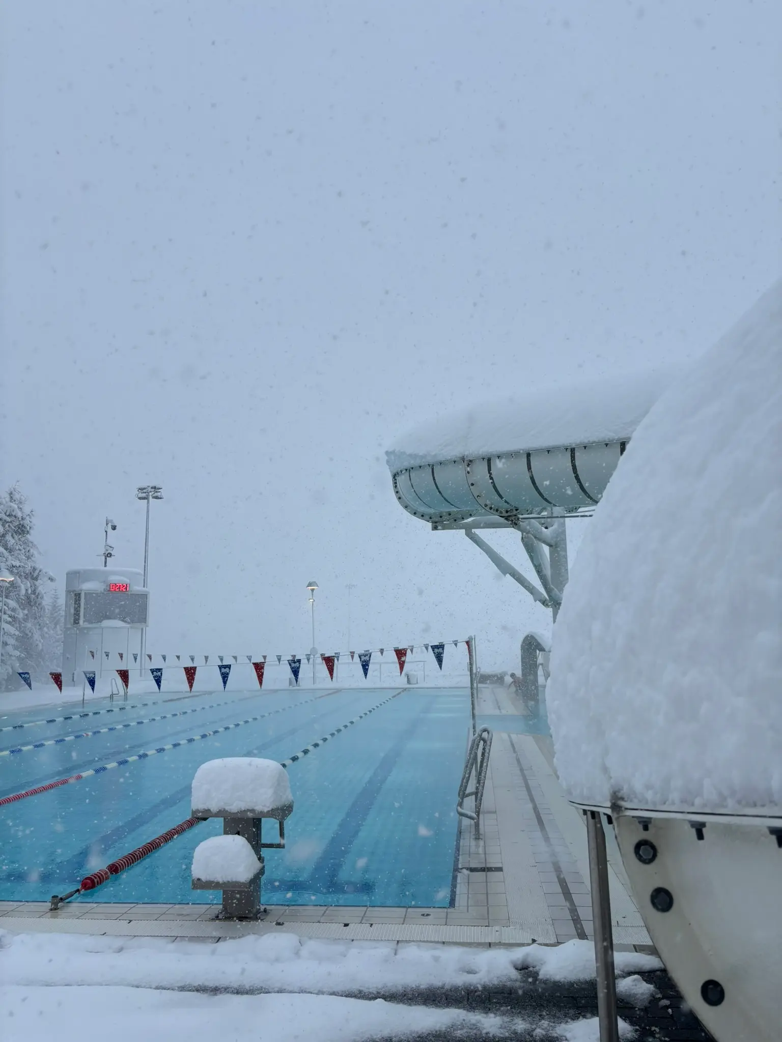 Outdoor swimming pool at Árbaejarlaug in Reykjavík during heavy snowfall, with snow-covered pool edges, lane dividers stretching into the distance, and a slide partially buried in fresh snow. The warm blue water contrasts with the white winter scene.
