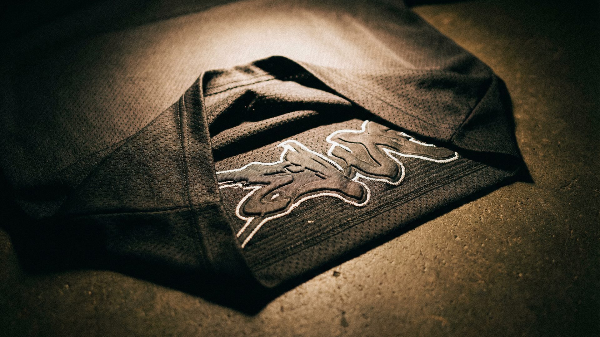 An extreme close-up of a custom hockey jersey laid flat on a dark raw concrete surface: the focal point is the layered embellishment detail showing a base layer of black twill fabric letters, a mid-layer of white embroidered stitching outlining the letters, and a top layer of raised puff print texture, all on a heavyweight charcoal mesh fabric with visible woven texture.