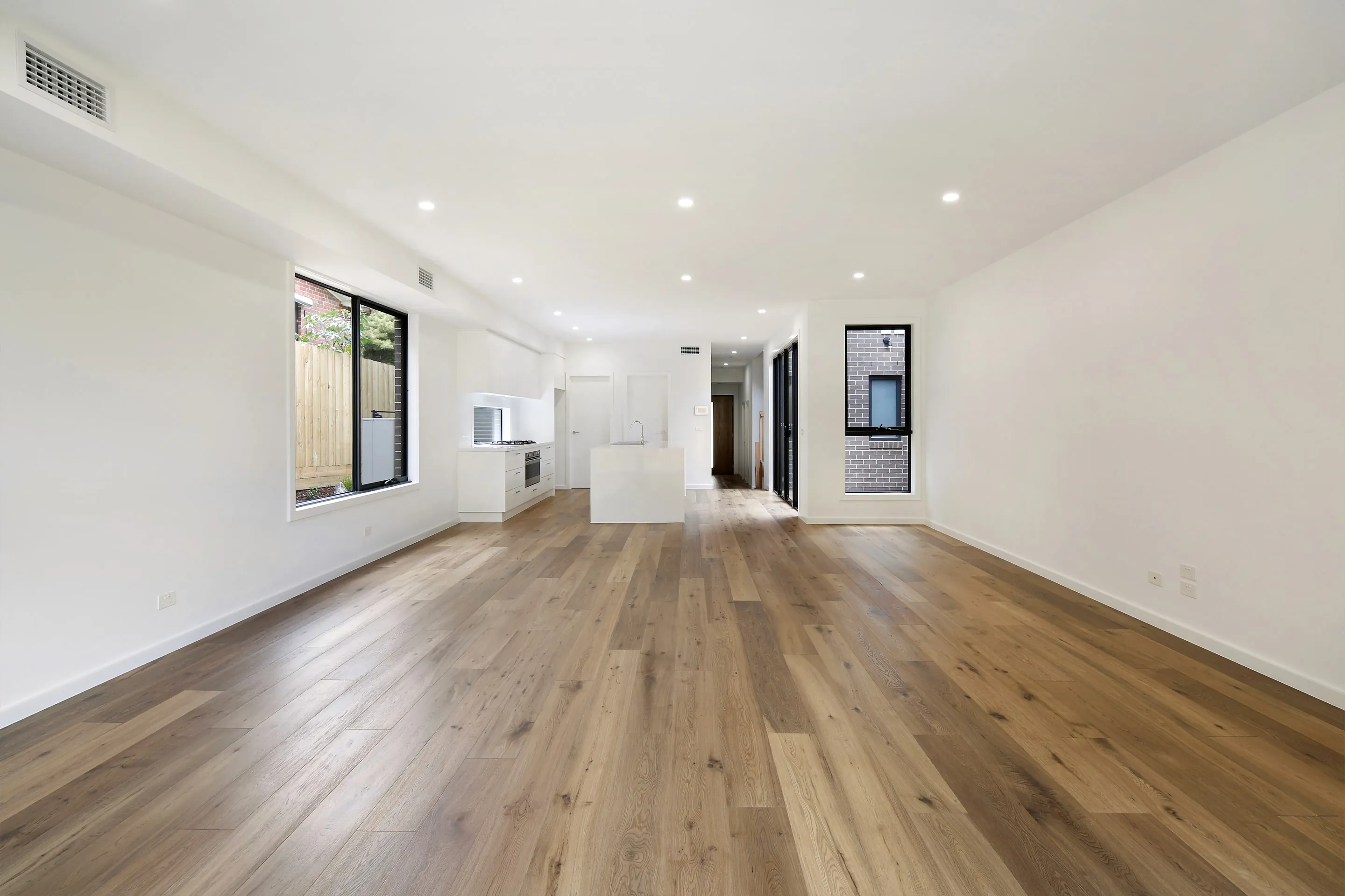Open living and kitchen with white walls, oak floors, a white island, black-framed windows to the garden and a brick courtyard, and a corridor leading to the stairs