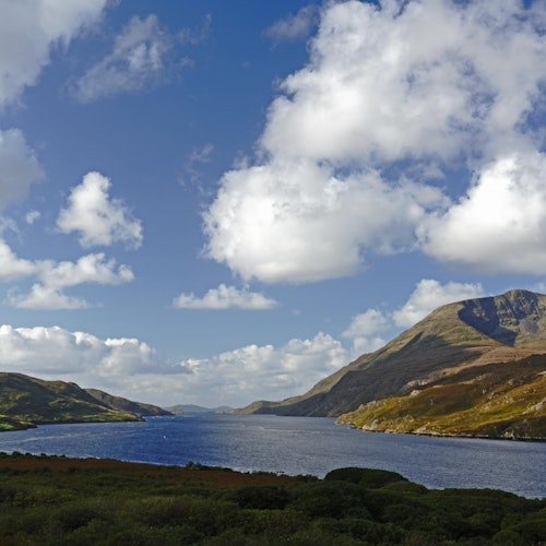 Спокойно езеро, заобиколено от зелени хълмове и планини под частично облачно синьо небе.