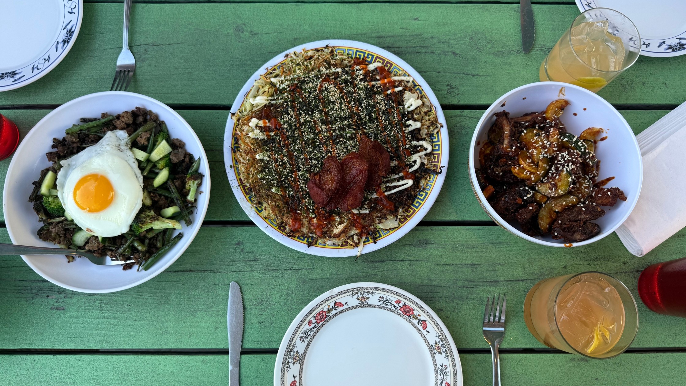 A trio of dishes from Xiao Bao Biscuit: Pad Kra Pow with spicy basil beef and a sunny side up egg, Okonomiyaki cabbage pancake topped with furikake mayo and sweet soy, and Som Tum with black bean fried chicken over rice and spicy green papaya salad.