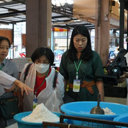 Tiga orang memerhatikan timbunan bahan putih dalam bekas biru di pasar tertutup, sementara seorang menunjuk dan seorang lagi merekod.