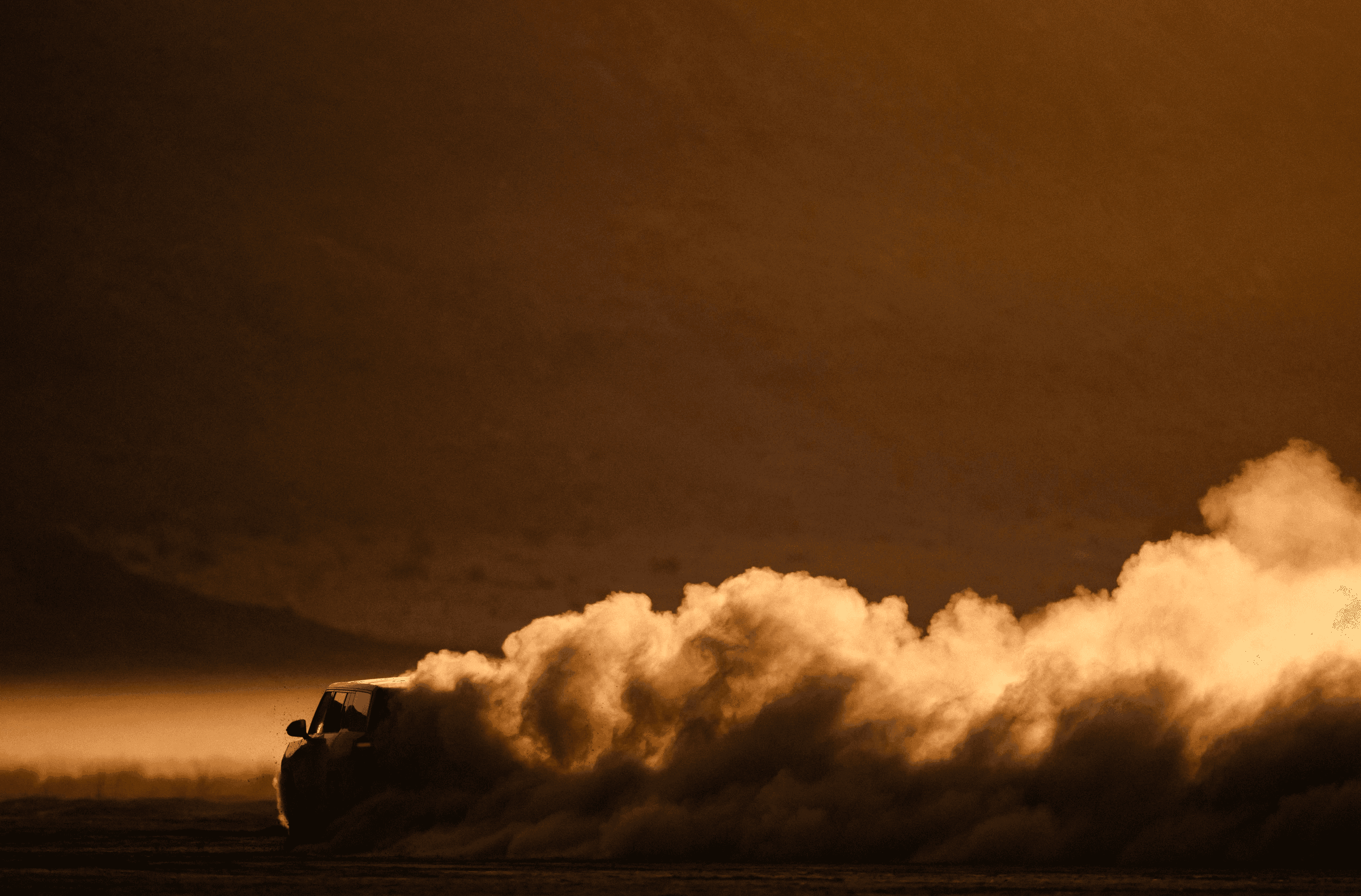 A Rivian R1S drives through a dusty landscape at sunset, kicking up a large cloud of dust behind it.