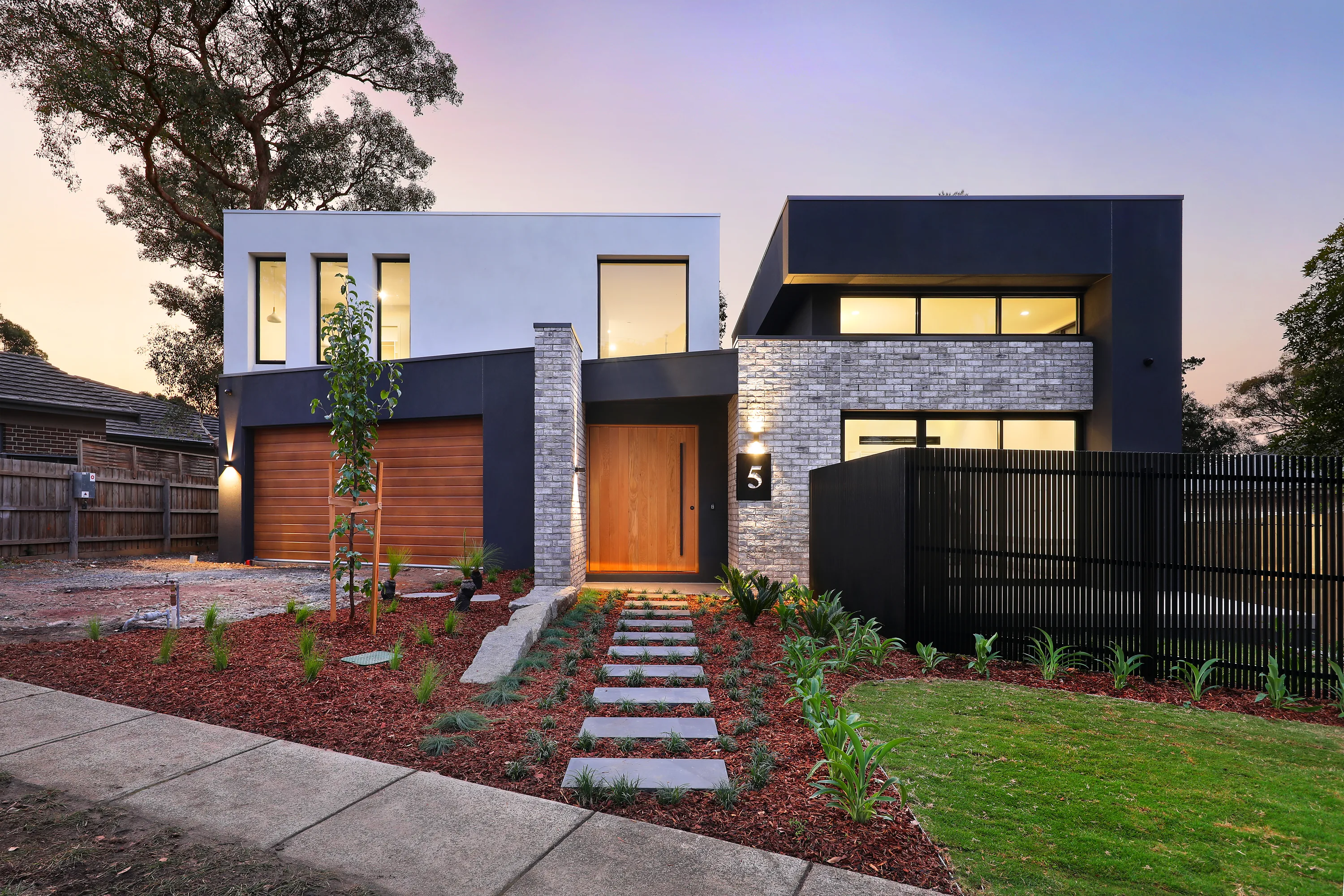 Front facade of the Montmorency Residence at dusk, a two-storey home pairing white render with a charcoal limewashed-brick wing, timber garage and entry door, house number 5 and a black batten fence