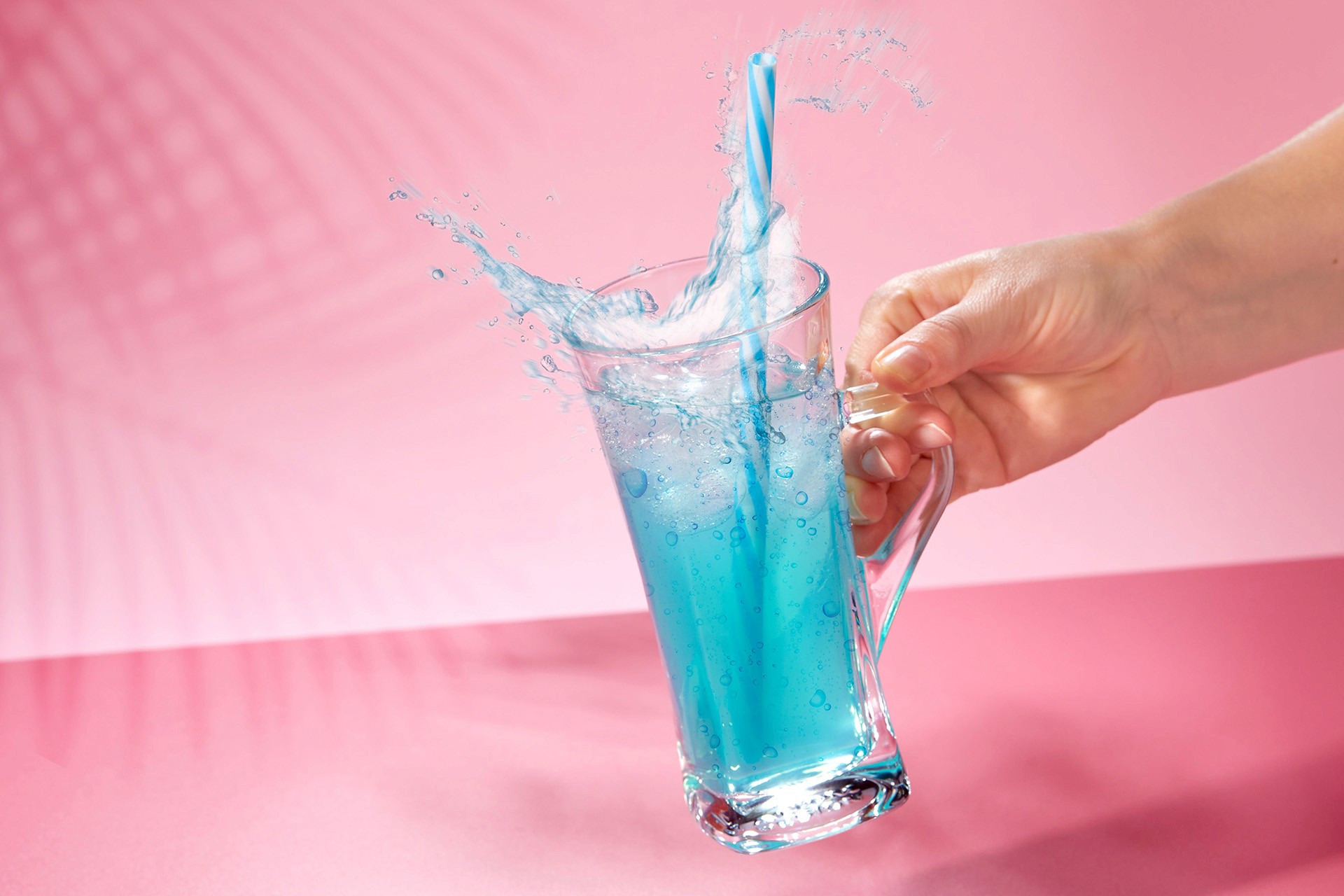 A hand holds a clear glass mug filled with vibrant blue liquid, splashing energetically against a soft pink background, creating a dynamic and refreshing visual contrast.