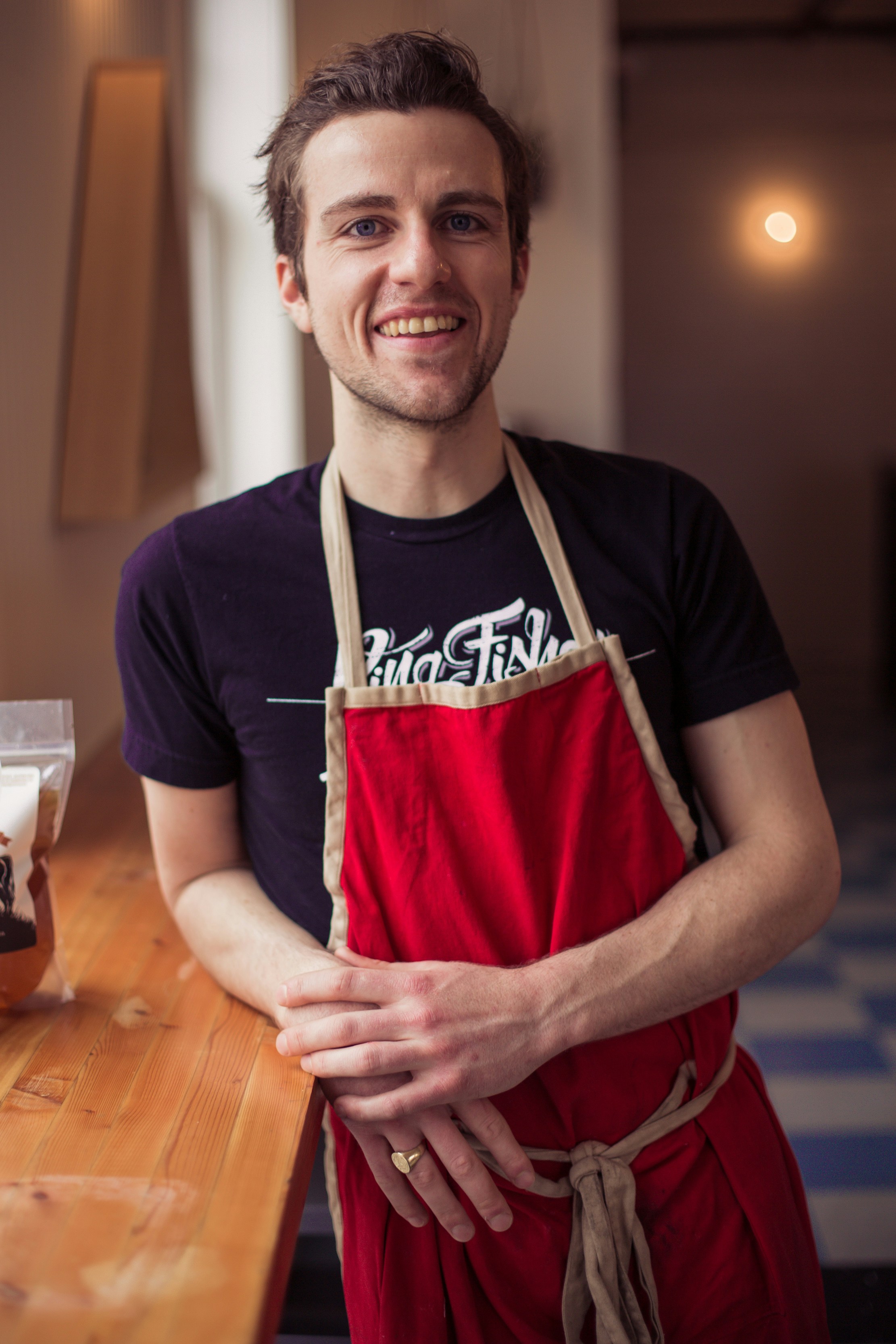 Smiling small business owner wearing a red apron and black t-shirt, ready to serve customers in his café or restaurant.