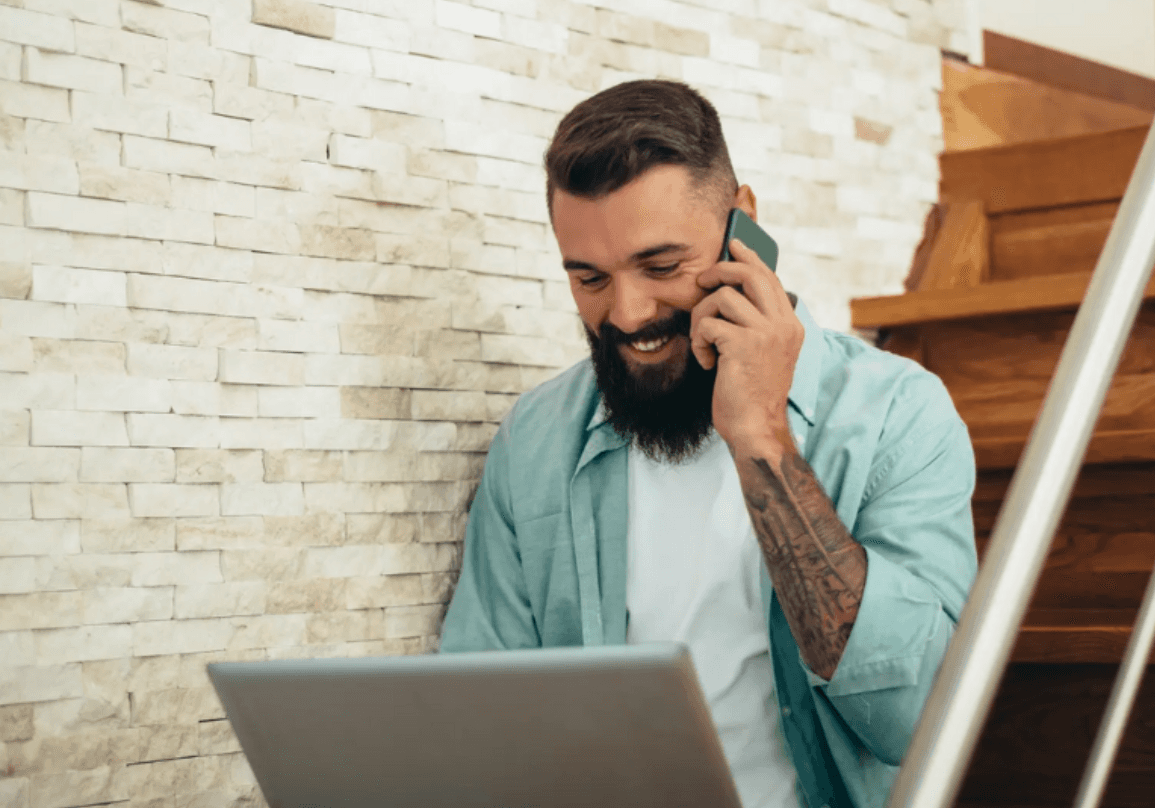 A smiling man sits on the stairs talking on the phone while on a laptop