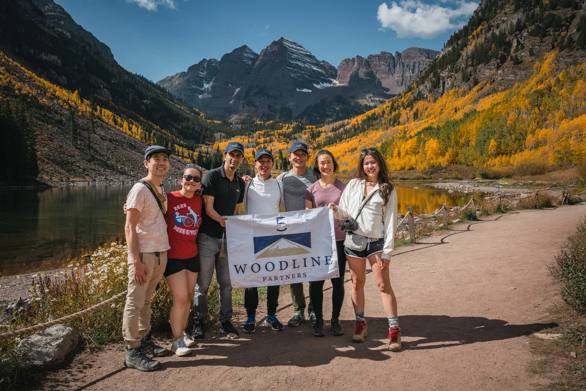Woodline Partners hiking group holds an anniversary banner beside Maroon Lake, with the Maroon Bells and golden aspens behind them.