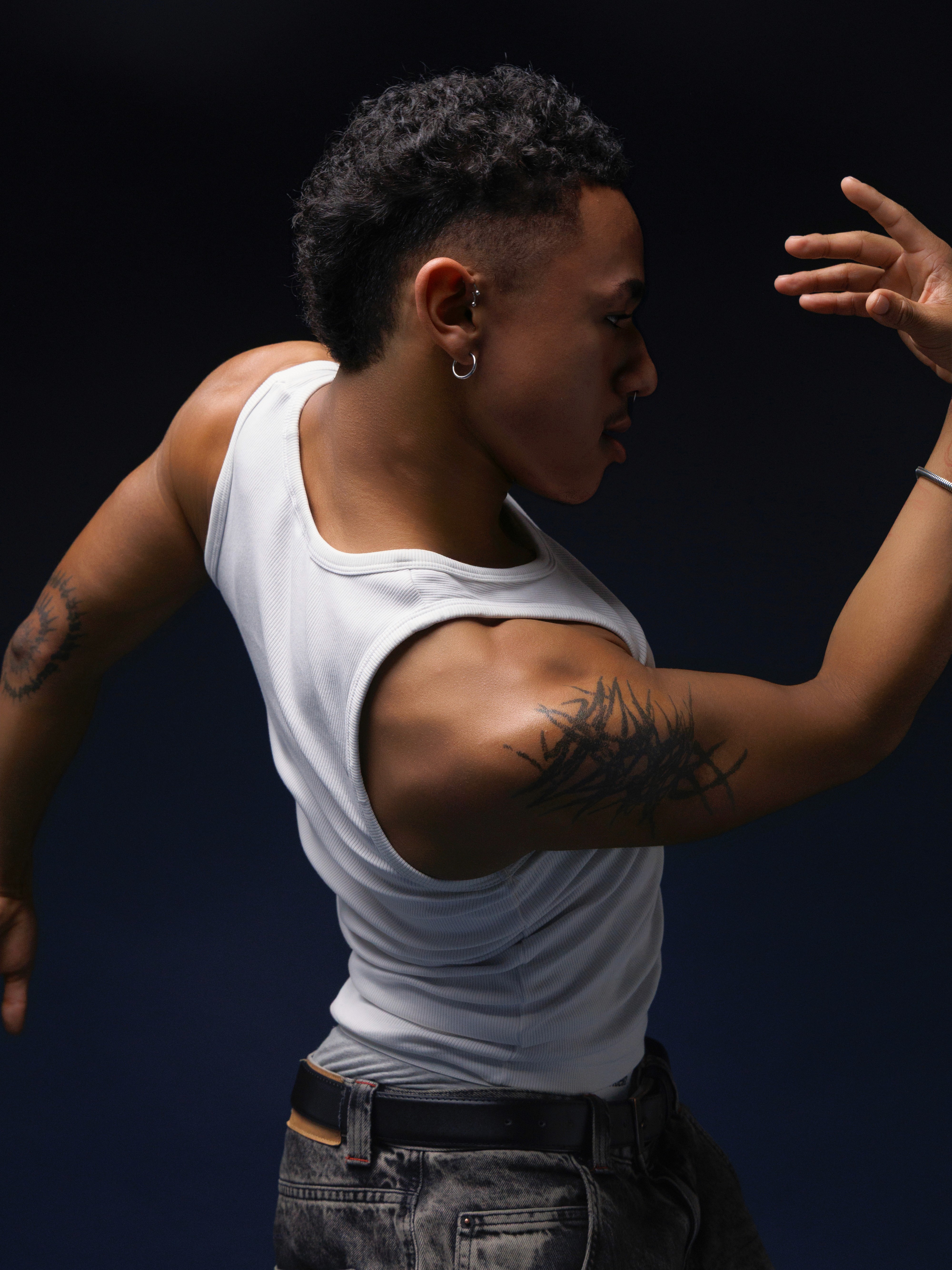 Boy in white tanktop posing with his hand pointing towards the sky and his body twisted, in front of a dark background.