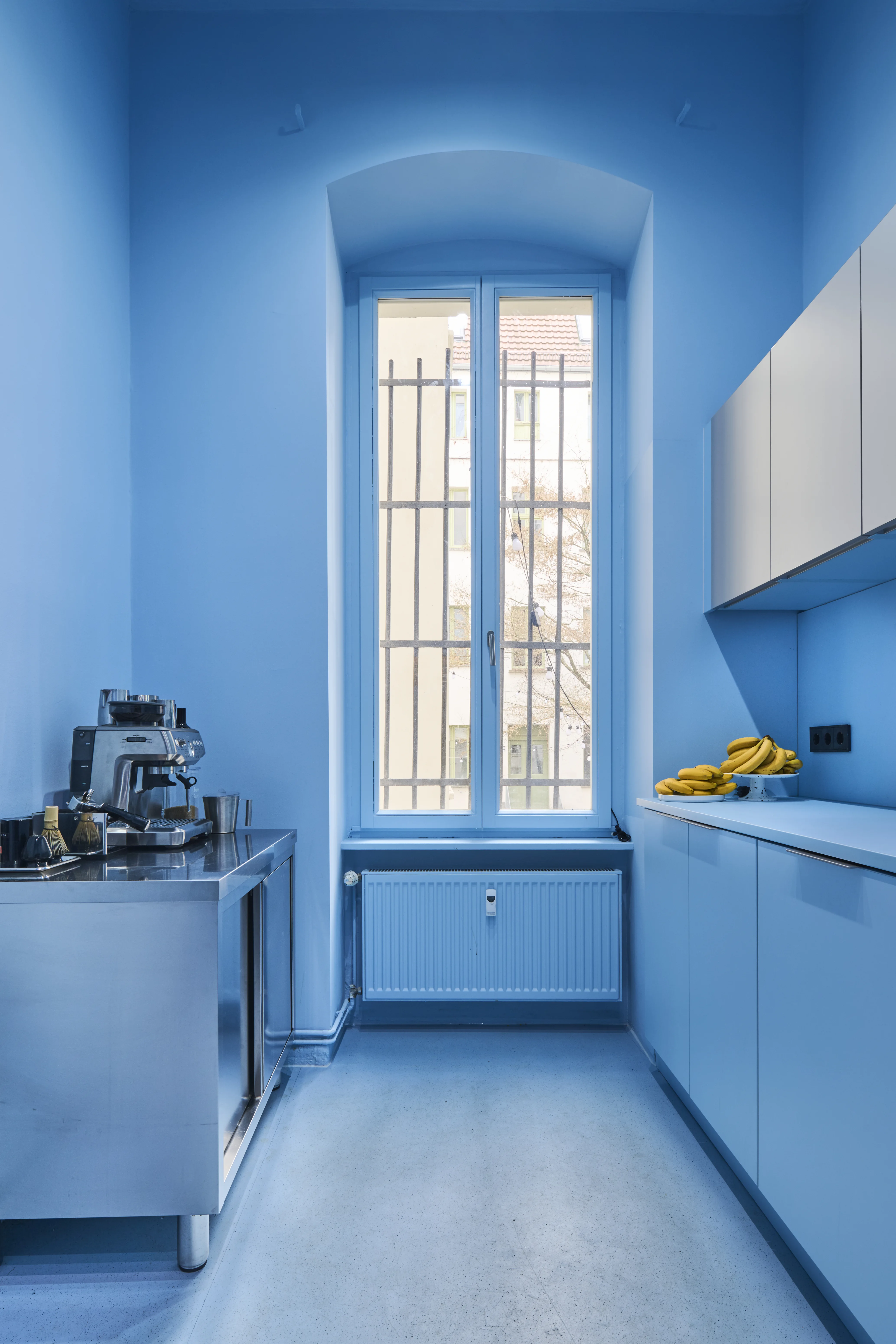 All-blue kitchen inside BOLD OFFICE with an arched barred window, a coffee machine on a stainless steel counter, bananas on the worktop, and white upper cabinets against blue walls.