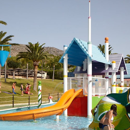 Barn leker i en färgglad vattenpark med rutschkanor och plaskfunktioner, omgivna av palmträd och grönområden.