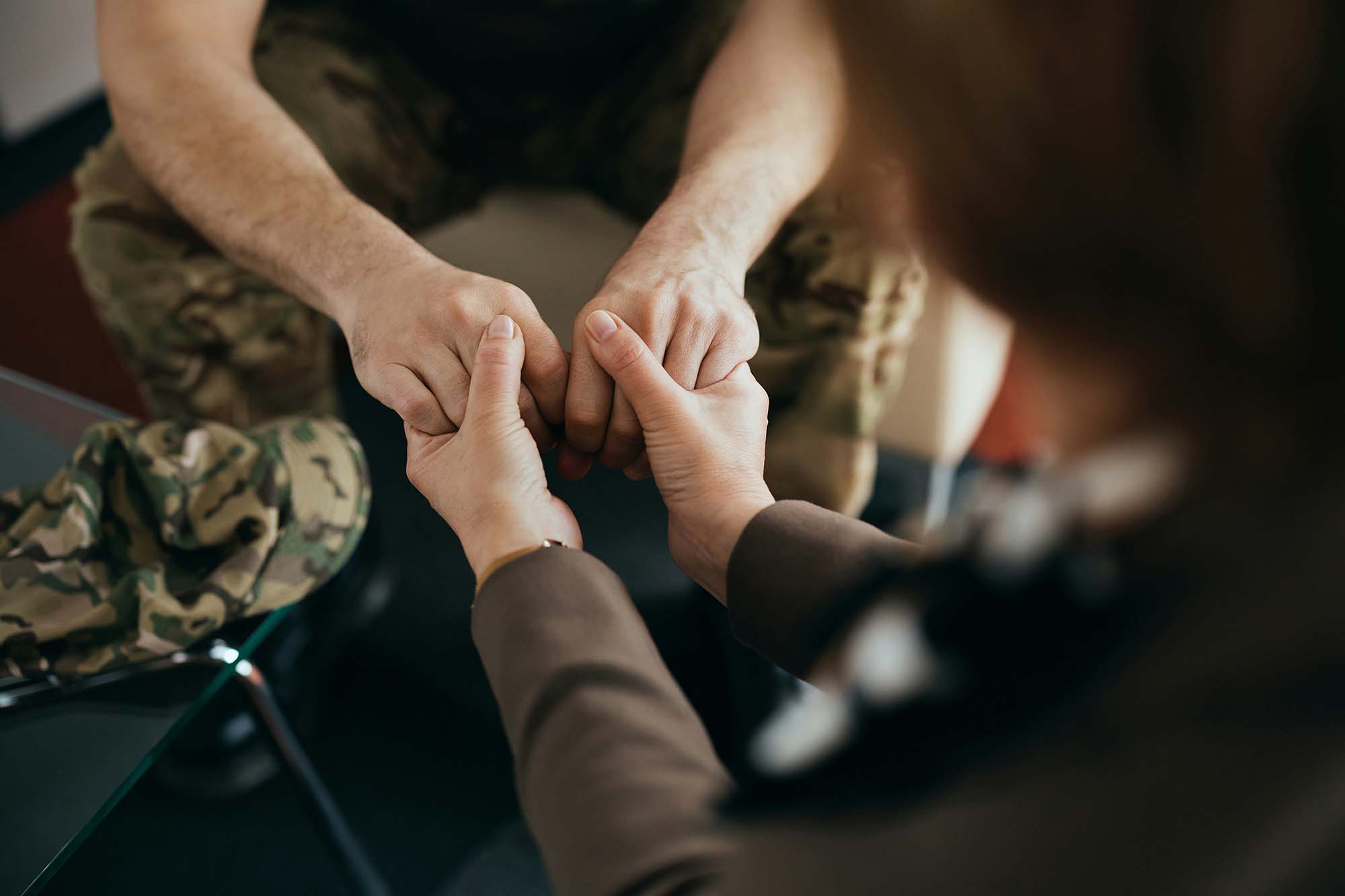 Two people holding hands. A man in military fatigues sits with hands clasped, another person's hands reach in from the bottom of the frame to clasp the man's hands.