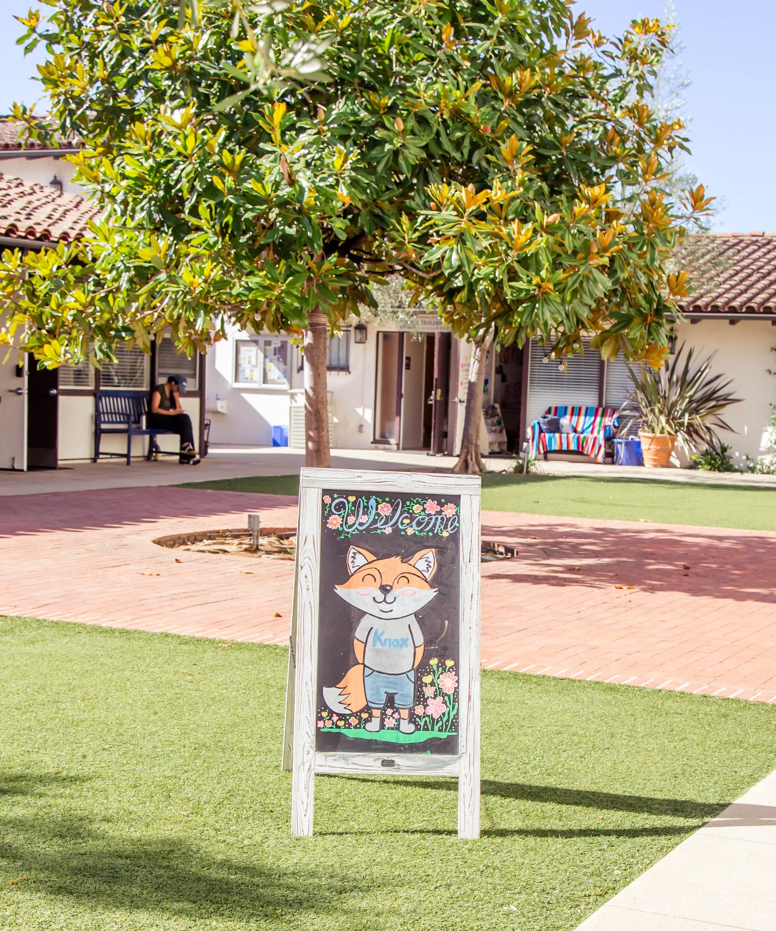 Welcome chalkboard sign with the Knox fox mascot on the lawn in front of the school buildings, beneath a magnolia tree