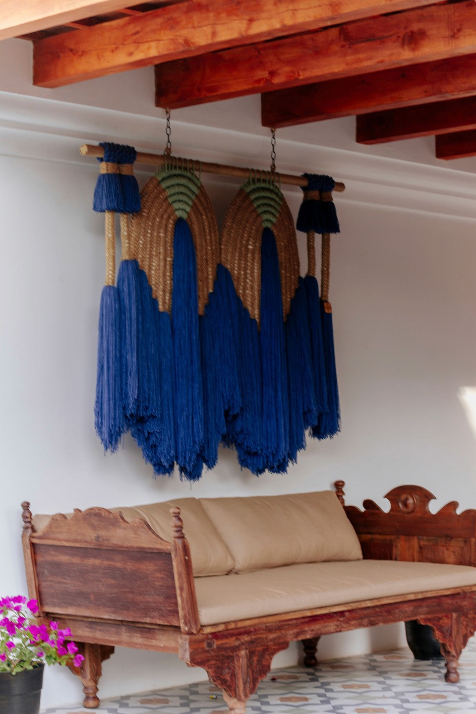 A rustic wooden bench with beige cushions sits against a white wall, below a large, decorative hanging featuring woven straw and vibrant blue tassels, adding an artistic flair to the space; a pot of pink flowers rests on the tiled floor nearby.