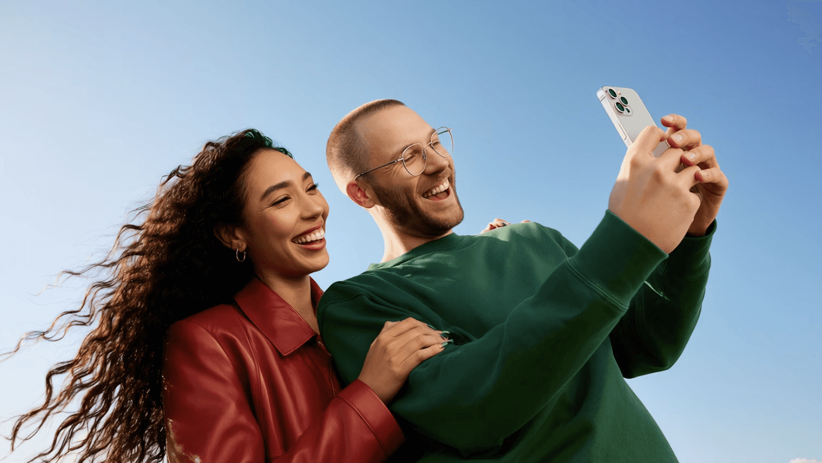 A cheerful couple takes a selfie outdoors under a clear blue sky, sharing a joyful moment together.