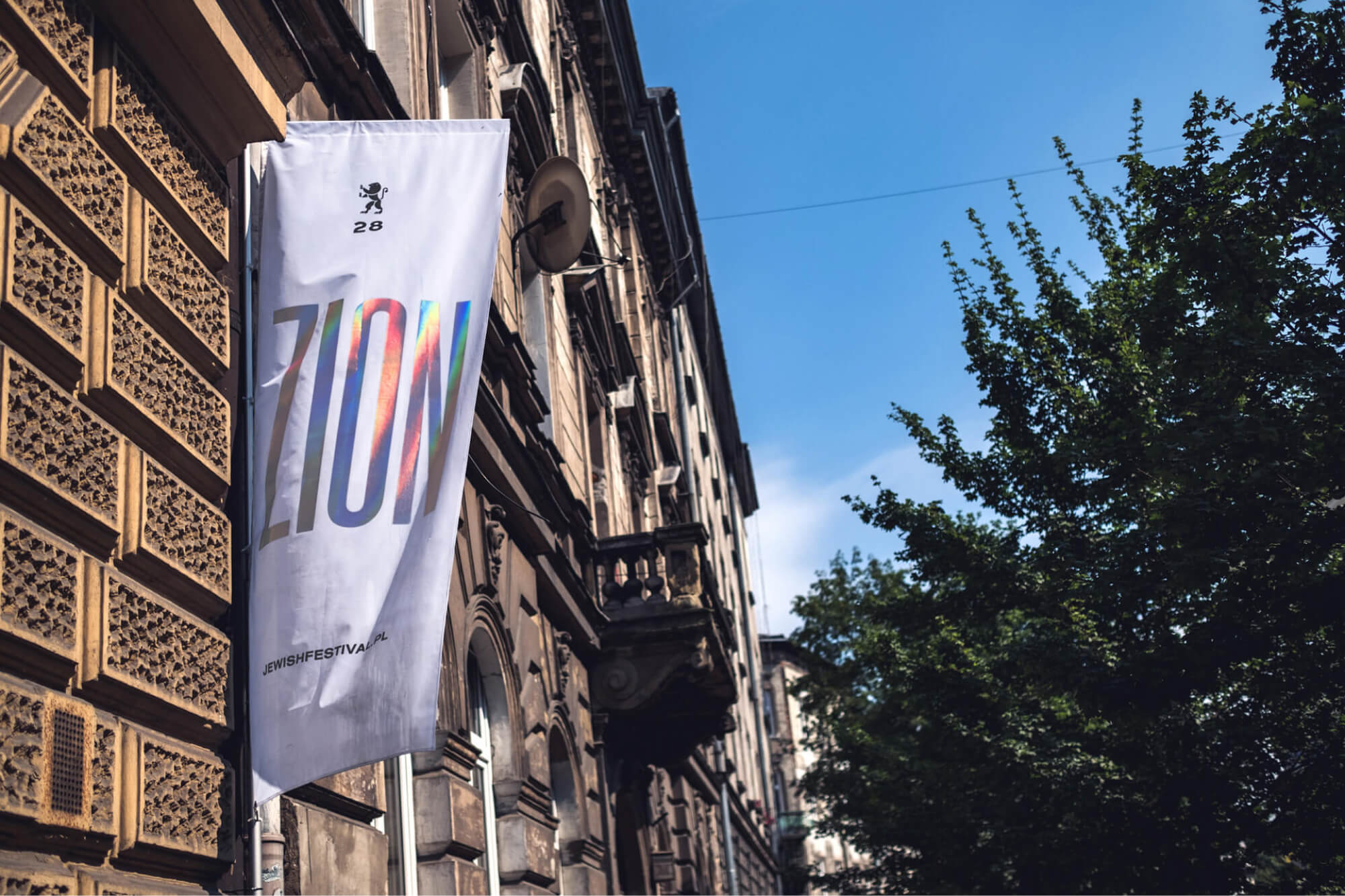 Biała pionowa flaga z gradientowym napisem „ZION” zwisa z ozdobnej fasady kamienicy nad ulicą.