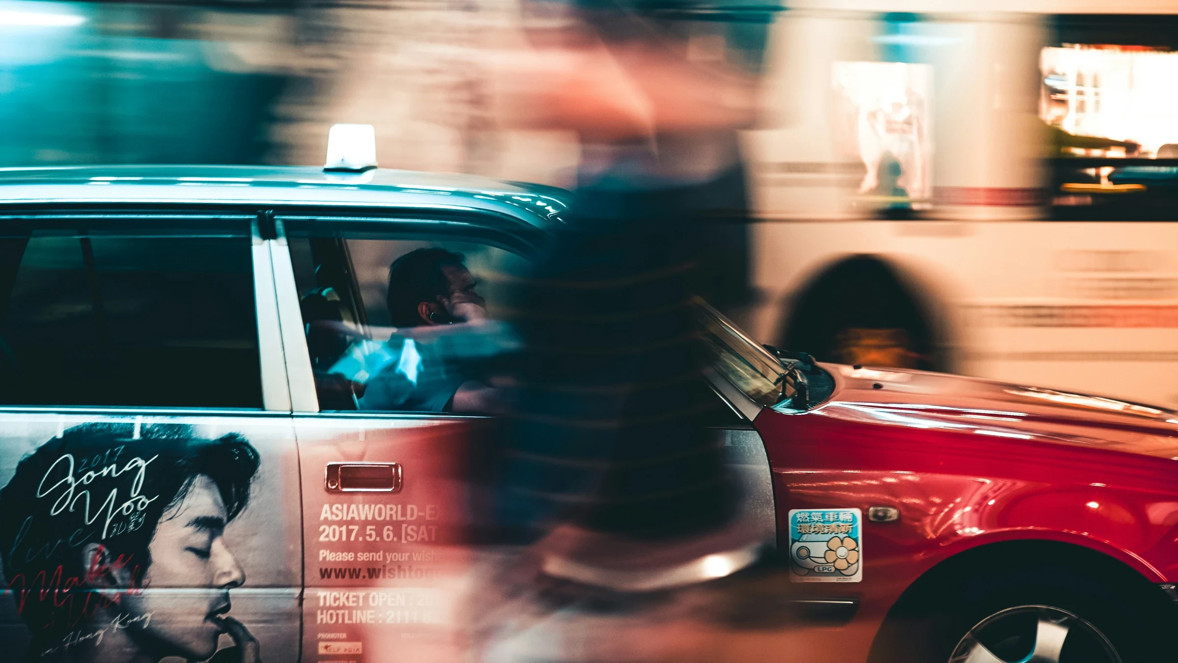 A red Hong Kong taxi on a busy street, with a passer-by captured in motion blur.