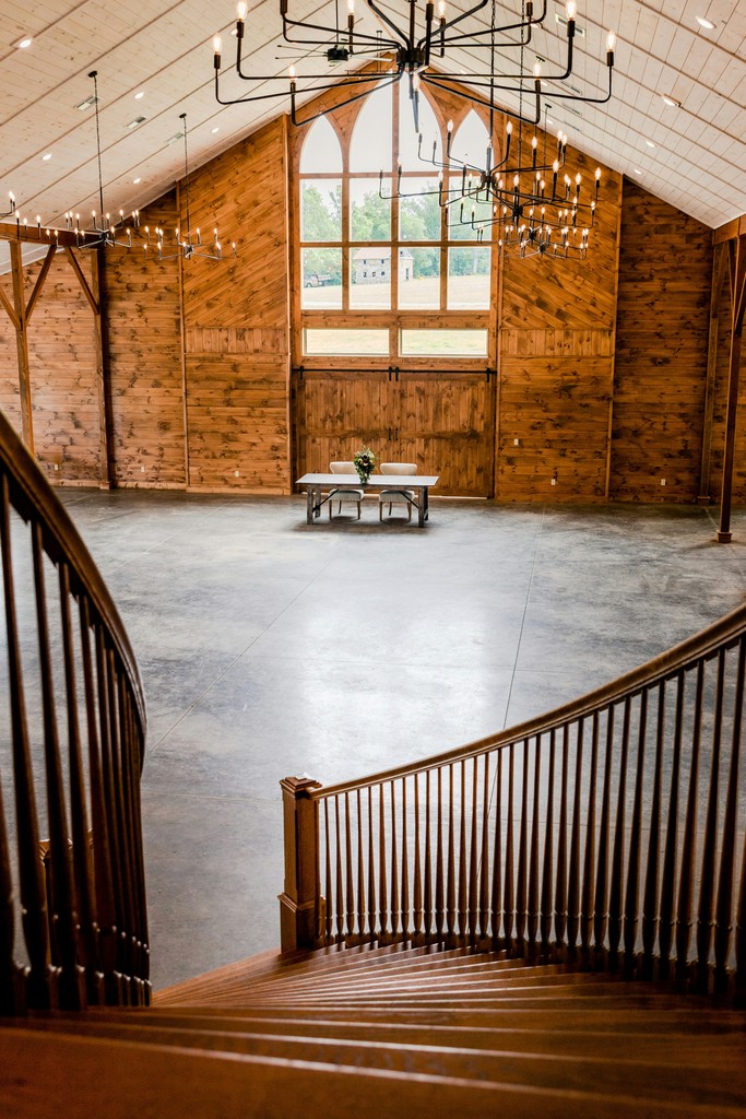 Elegant barn wedding venue interior with vaulted wooden ceilings, modern chandeliers, and a curved staircase overlooking the reception floor