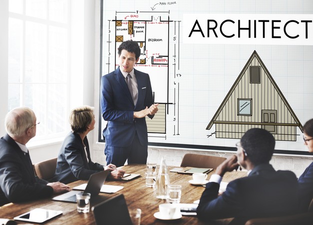 A man in a suit presents to a group at a table, with an "ARCHITECT" sign and a caution symbol displayed behind him.