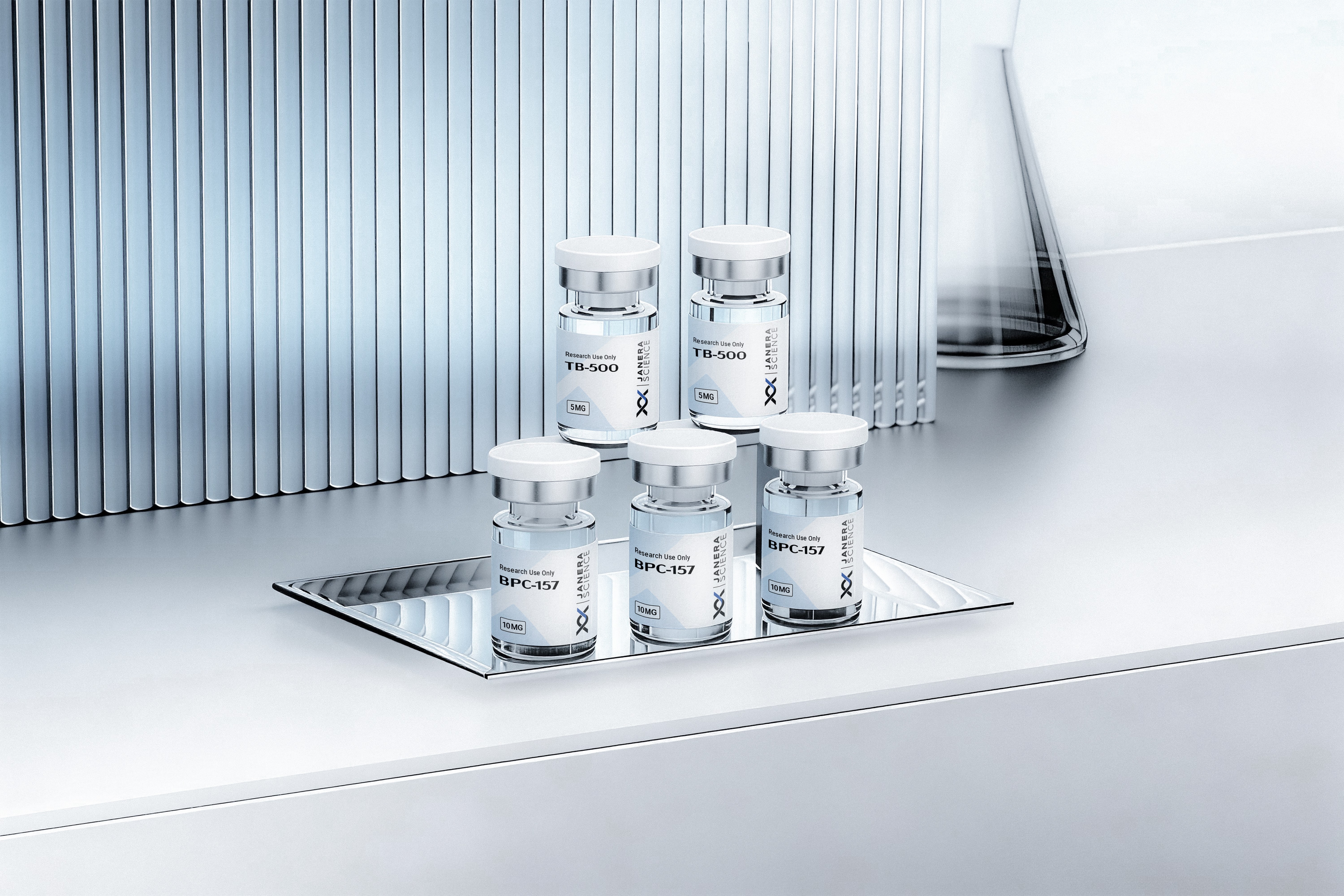Five Janera Science research peptide vials — two TB-500 5mg and three BPC-157 10mg — arranged on a mirrored stainless steel tray against a silver ridged laboratory background. For research use only.