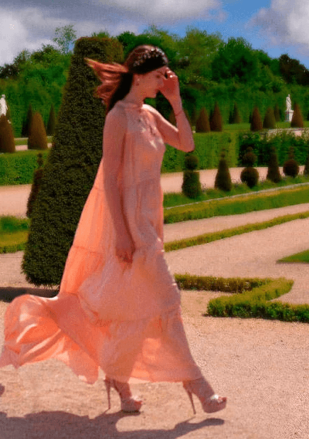 A woman in a flowing peach dress and high heels walks gracefully through a manicured Louvre garden, with neatly trimmed hedges and lush greenery under a partly cloudy sky, embodying elegance and natural beauty in Abarel's photo session.