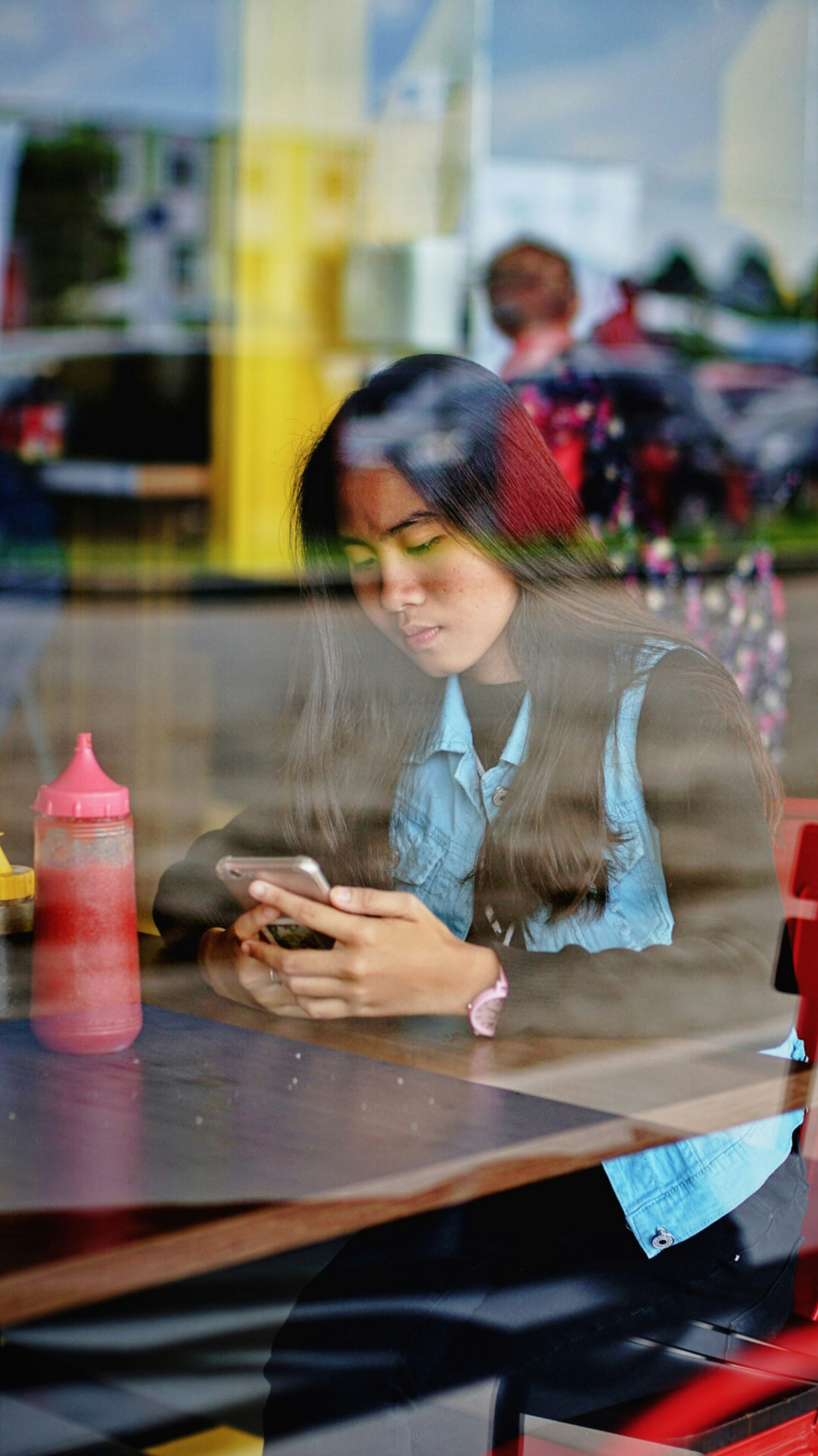 A young woman with long hair sits in a cafe looking at her phone, photo is taken through the glass