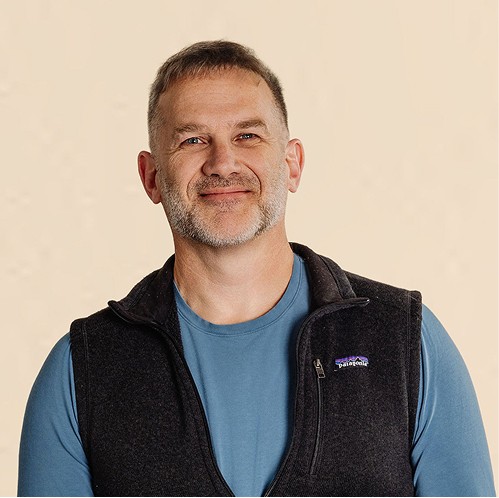 Man with short hair and light stubble wearing a blue shirt and dark vest, standing against a neutral background.