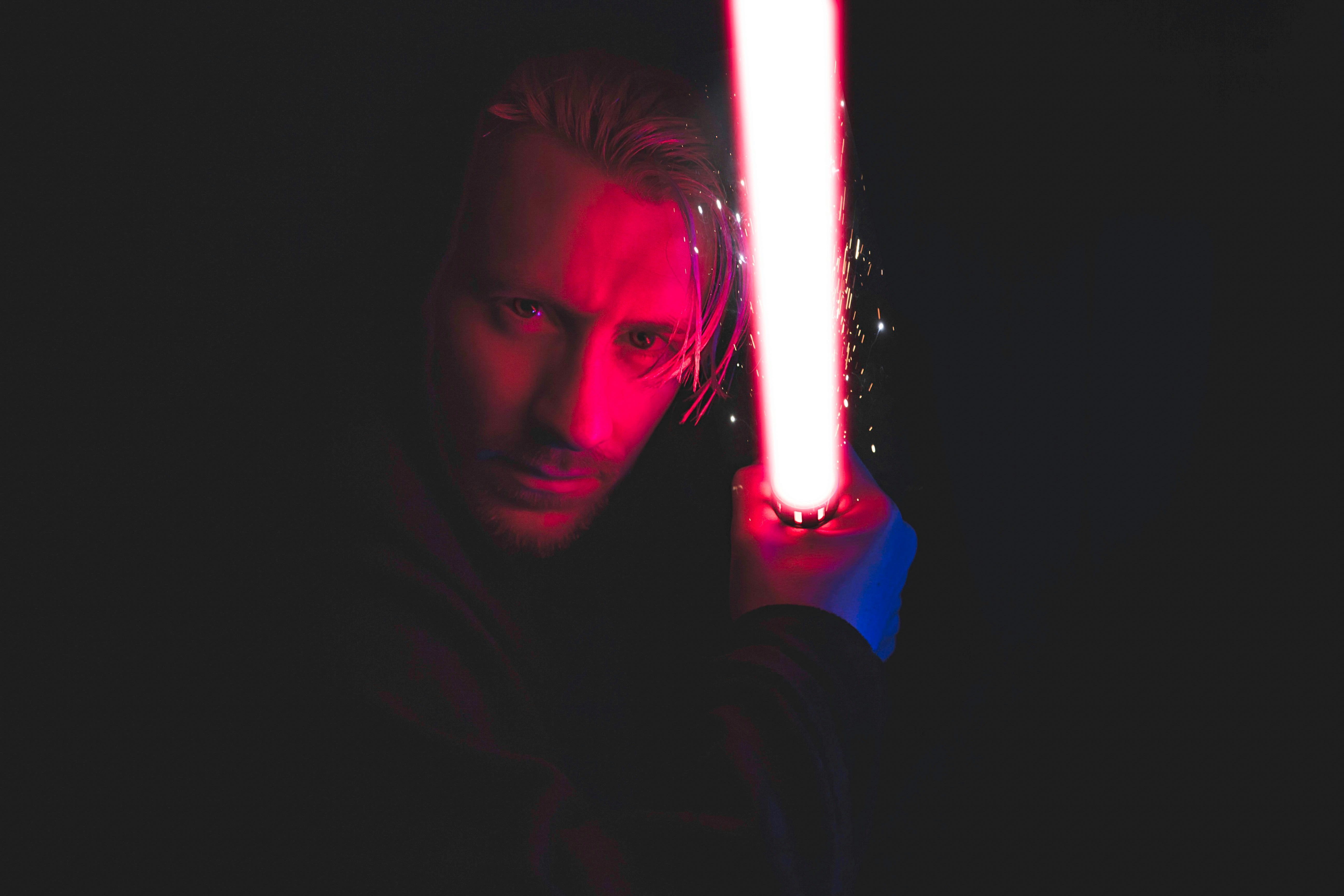 A person holding a glowing red lightsabre in a dark environment. The face is partially illuminated by the red light, creating a dramatic effect. The background is mostly black, enhancing the focus on the lightsaber and the person's intense expression.